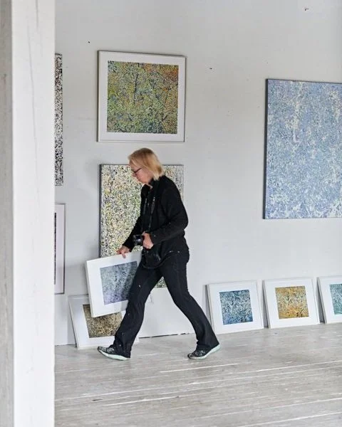 A person in a black outfit carrying a framed piece of art in an art gallery with abstract paintings on the walls.