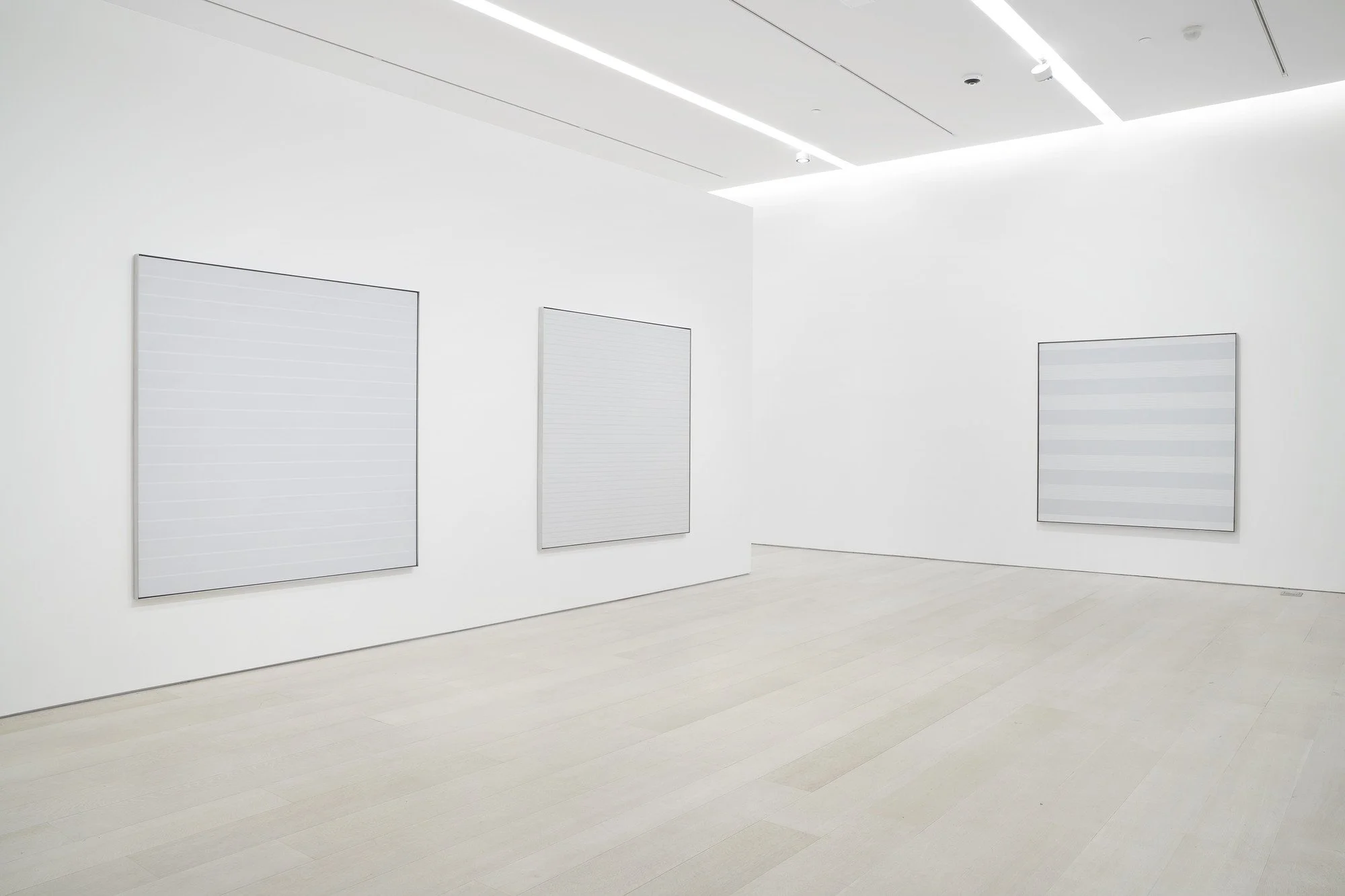A white gallery space with three large square paintings with faint grids on them. Paintings by Agnes Martin. Pace Gallery.