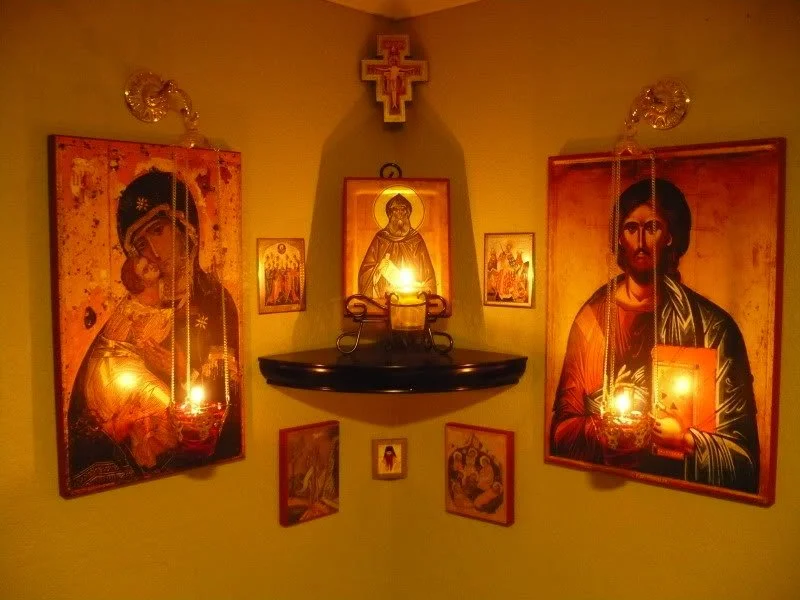 Traditional Russian icon corner with Orthodox images of the Virgin and Child, Christ, and saints arranged where two walls meet, illuminated by small lamps and candles.