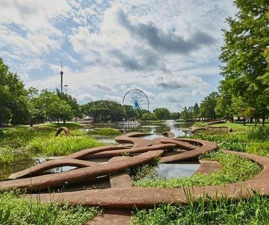 Patricia Johanson’s Fair Park Lagoon at Fair Park in Dallas, with reddish-brown concrete walkways looping through shallow water and aquatic plants. Trees and a Ferris wheel appear in the background.