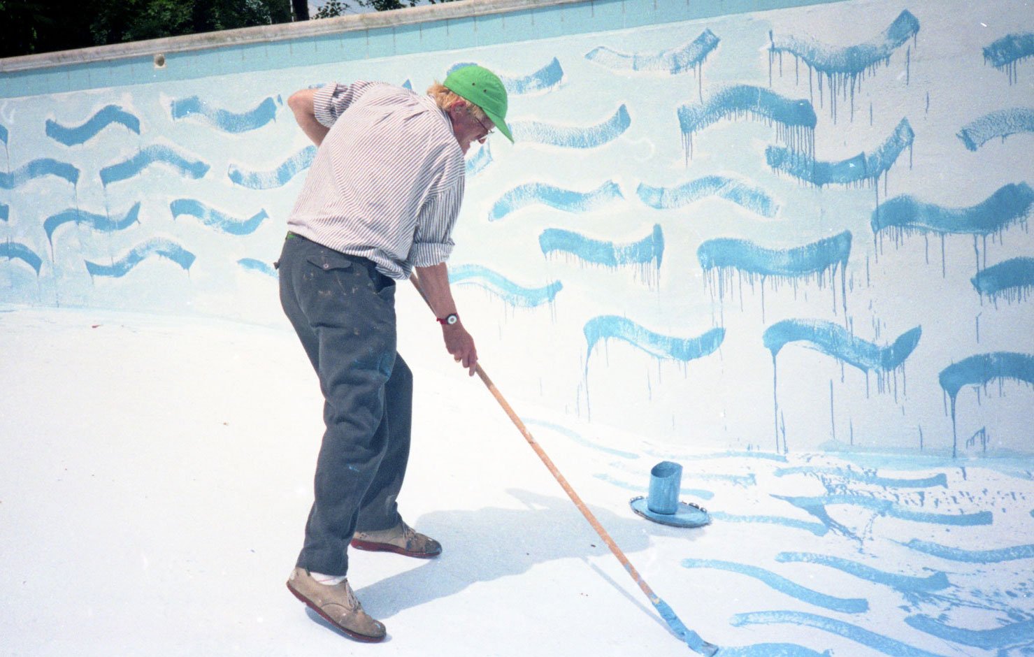 David Hockney painting the interior of an empty swimming pool at Ken Tyler’s home, using a long-handled brush to cover the pale blue surface with loose, wave-like blue marks. Hockney wears a green cap and striped shirt as he works inside the pool.