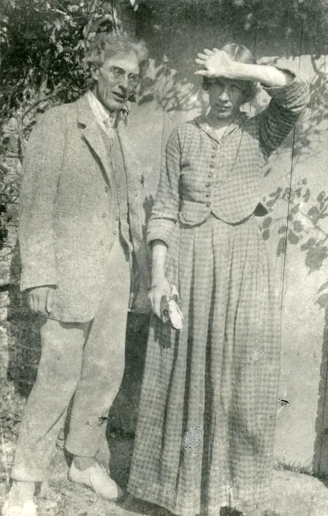 Black-and-white photograph of two adults standing outdoors against a sunlit wall. A man in a light suit stands beside a woman in a long patterned dress and vest, raising her hand to shield her eyes from the light.