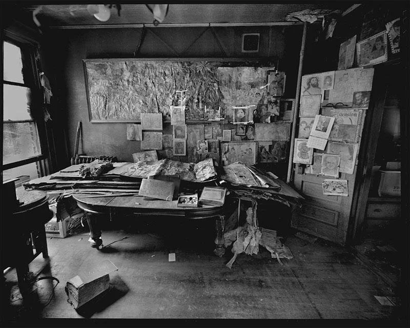 Black-and-white photograph of Henry Darger’s cluttered Chicago room and workspace, filled with stacks of drawings, papers, collage materials, and images pinned densely across the walls.