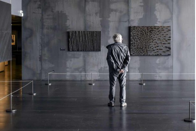 Textured black paintings hanging on a charcoal gray wall that is also textured, with a man standing in front of the painting in a gray jacket and pants. He has white hair, seen from behind.