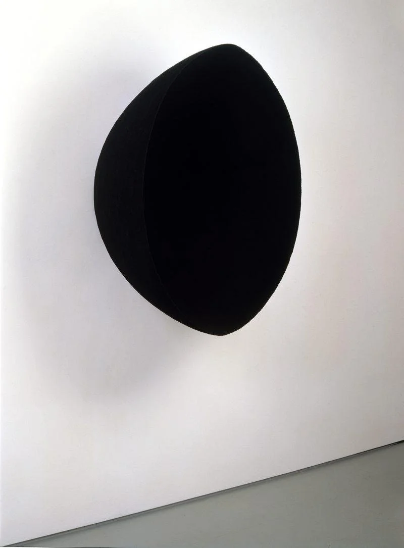 A oval shape hangs suspended on a wall, but you cannot be sure of its shape because it is so black you cannot tell if it is flat shape or something three dimensional.