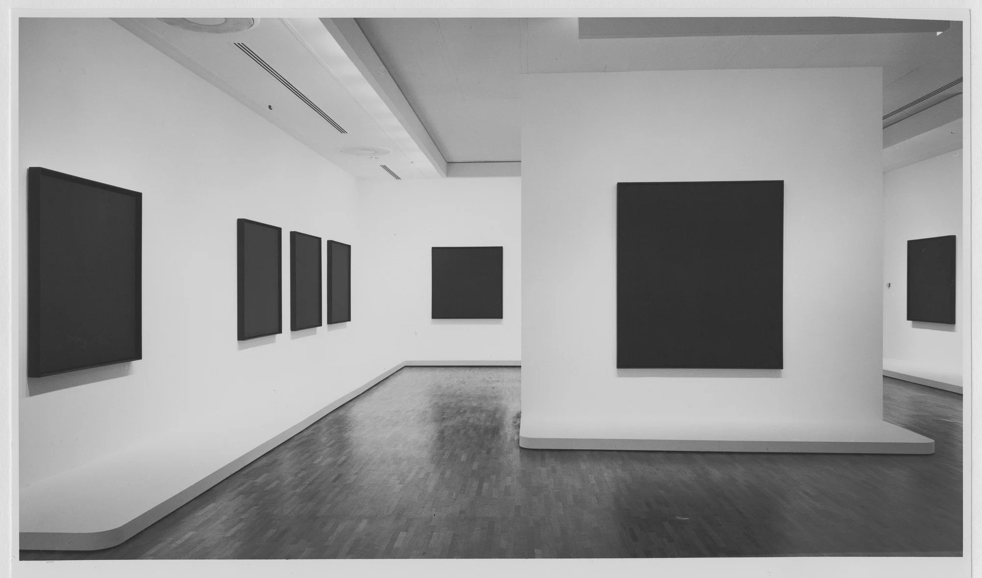 Seven all black paintings by Ad Reinhardt hung on white walls in the Museum of Modern art.