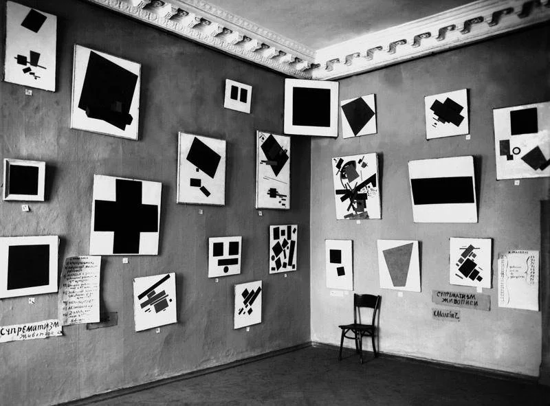 Black-and-white installation photograph of Malevich’s Suprematist paintings at the 1915 0.10 exhibition in Petrograd, with Black Square hung high in the corner of the room.