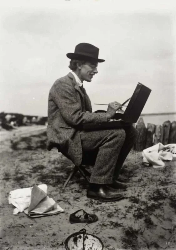 Black-and-white photograph of a man seated outdoors on a folding stool, painting at a portable easel. He wears a suit and hat, holds brushes in one hand, and works intently amid scattered papers on the ground.