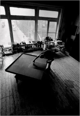 Ad Reinhardt painting in his New York studio, standing over a black painting with brush in hand, light coming into the studio through a large window.