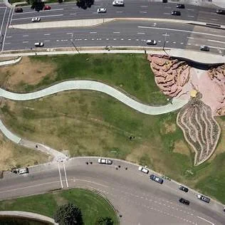 Aerial view of Patricia Johanson’s The Draw at Sugar House in Salt Lake City, with a pale curving pedestrian path, a sculptural Sego Lily form, and red canyon-like structures integrated into a flood-control landscape beside a roadway.