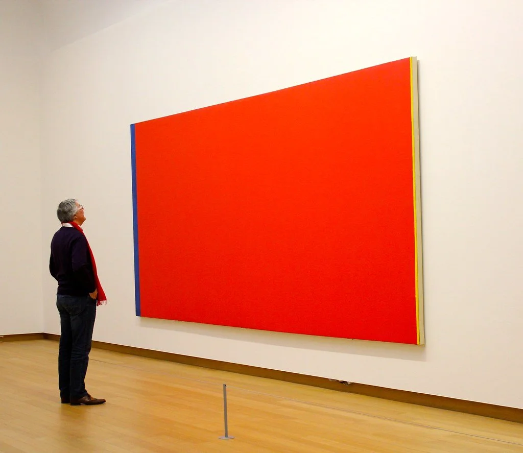 Visitor viewing Barnett Newman's Be I (1949), a monumental red color field painting framed by narrow vertical zips at either side.