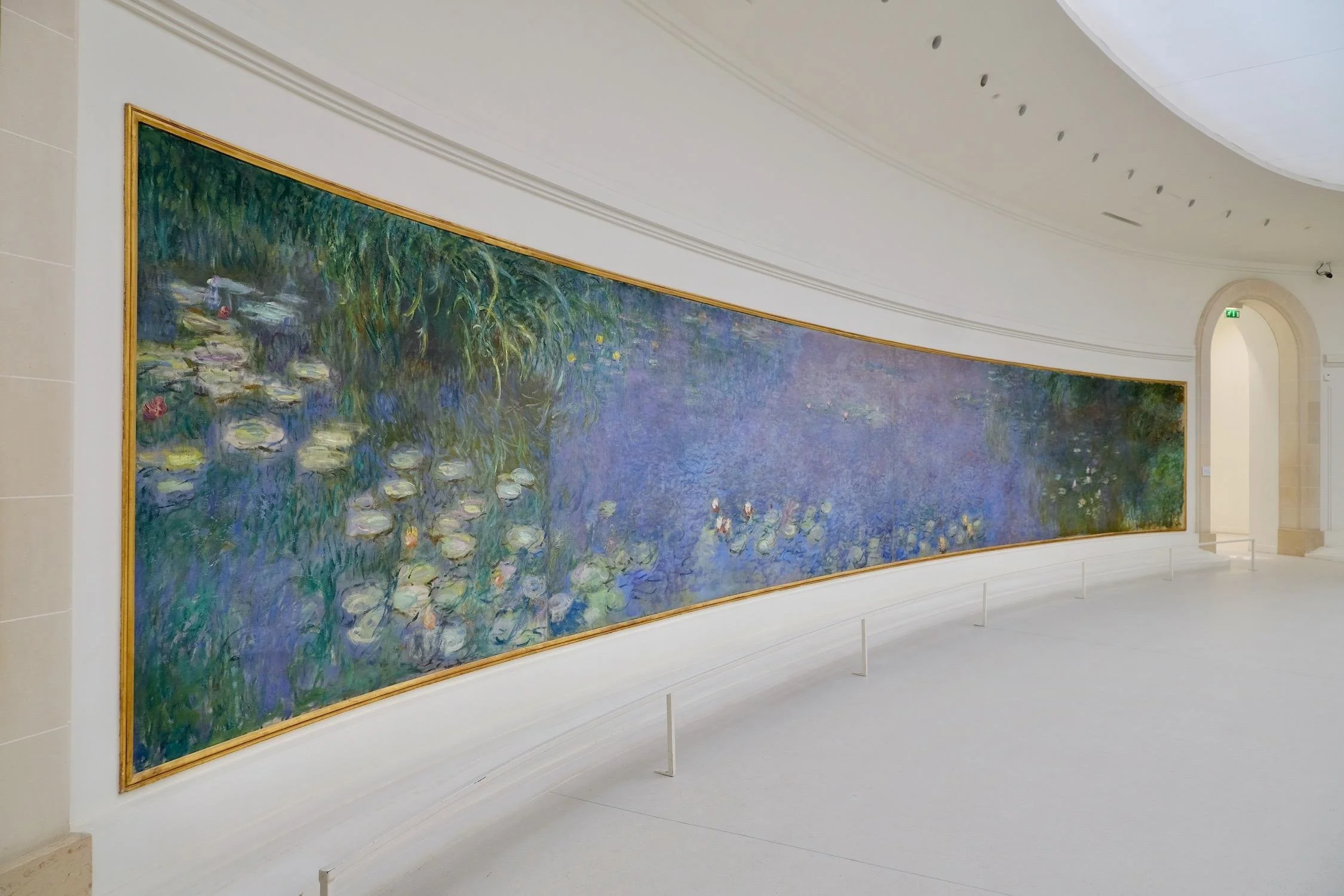 Claude Monet’s panoramic Water Lilies installation at the Musée de l’Orangerie in Paris. A vast curved canvas stretches across a white oval gallery, filled with shimmering blues, greens, violets, and floating lily pads.