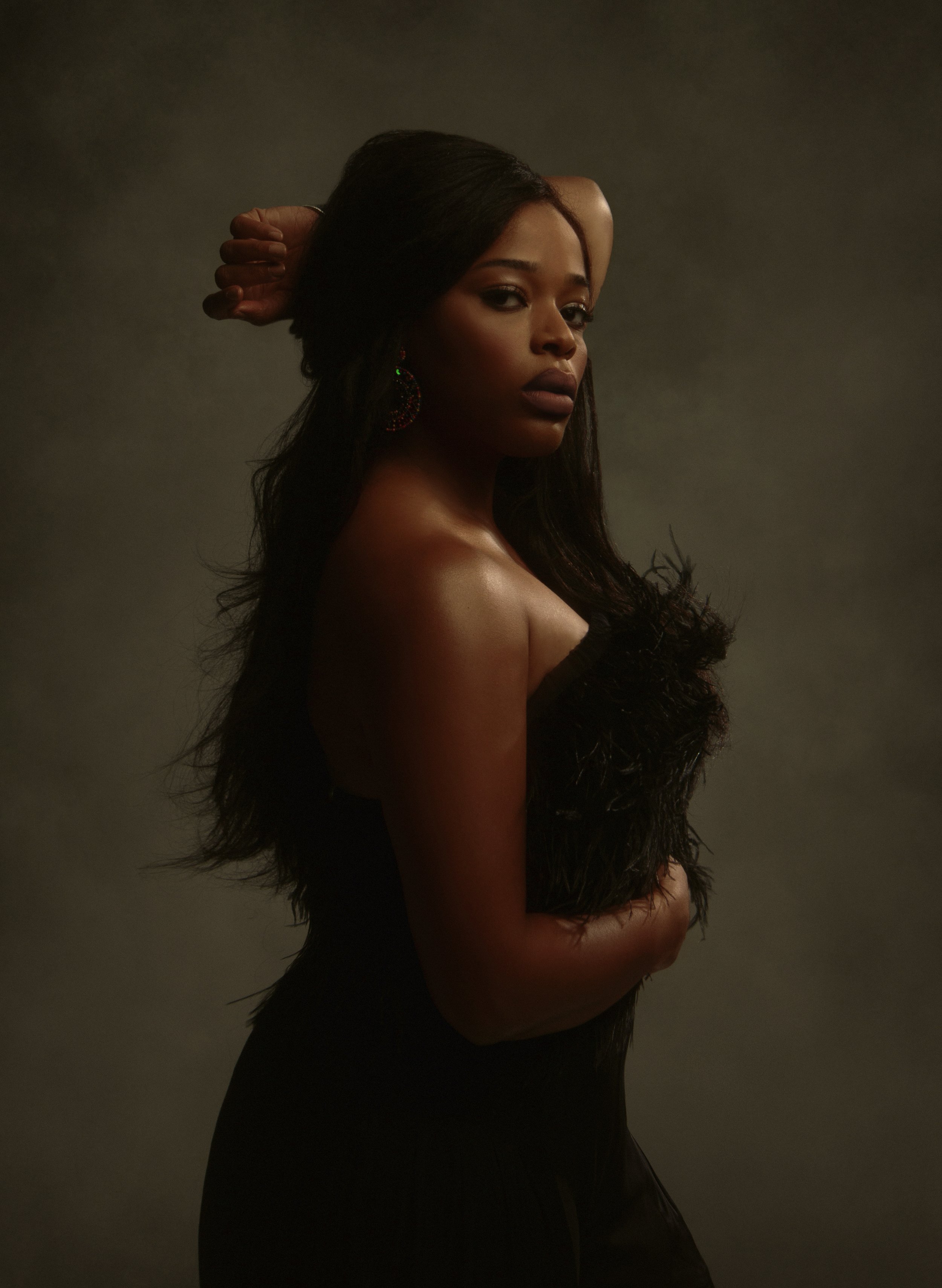 A young woman with dark hair and dark skin, posing with her hand behind her head and wearing a black fur-like strapless top and large earrings, against a dark gradient background.