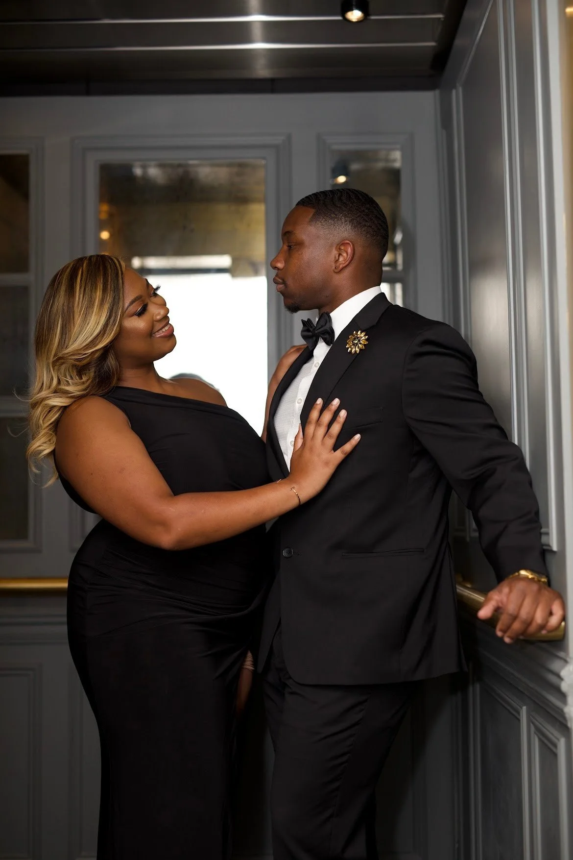 A couple dressed in formal attire, with the woman in a black gown and the man in a tuxedo, standing close together and looking at each other.
