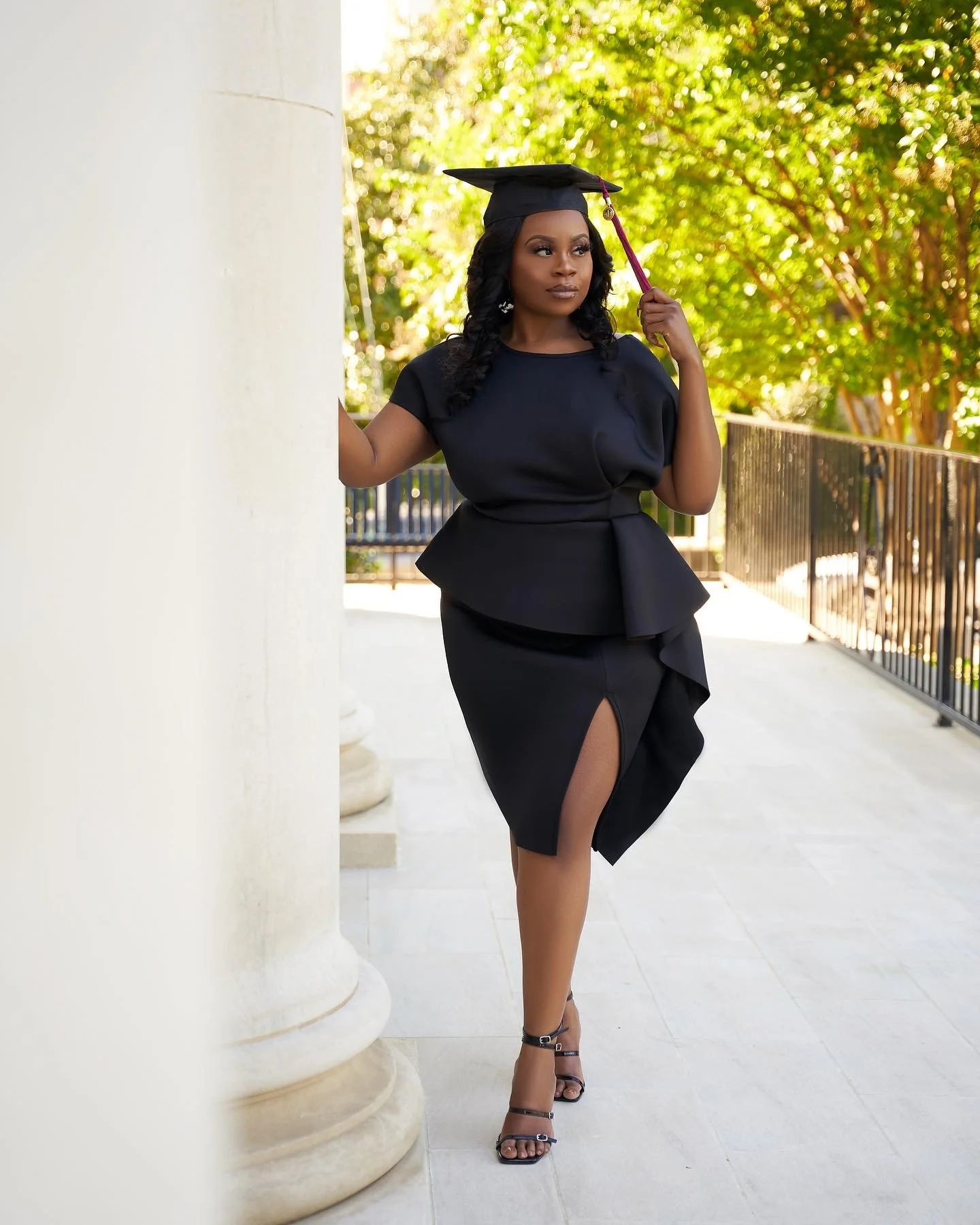 A woman in a black dress and heels standing outdoors near a white pillar, wearing a graduation cap. She is holding the cap's tassel with one hand, with a background of trees and a sunny atmosphere.