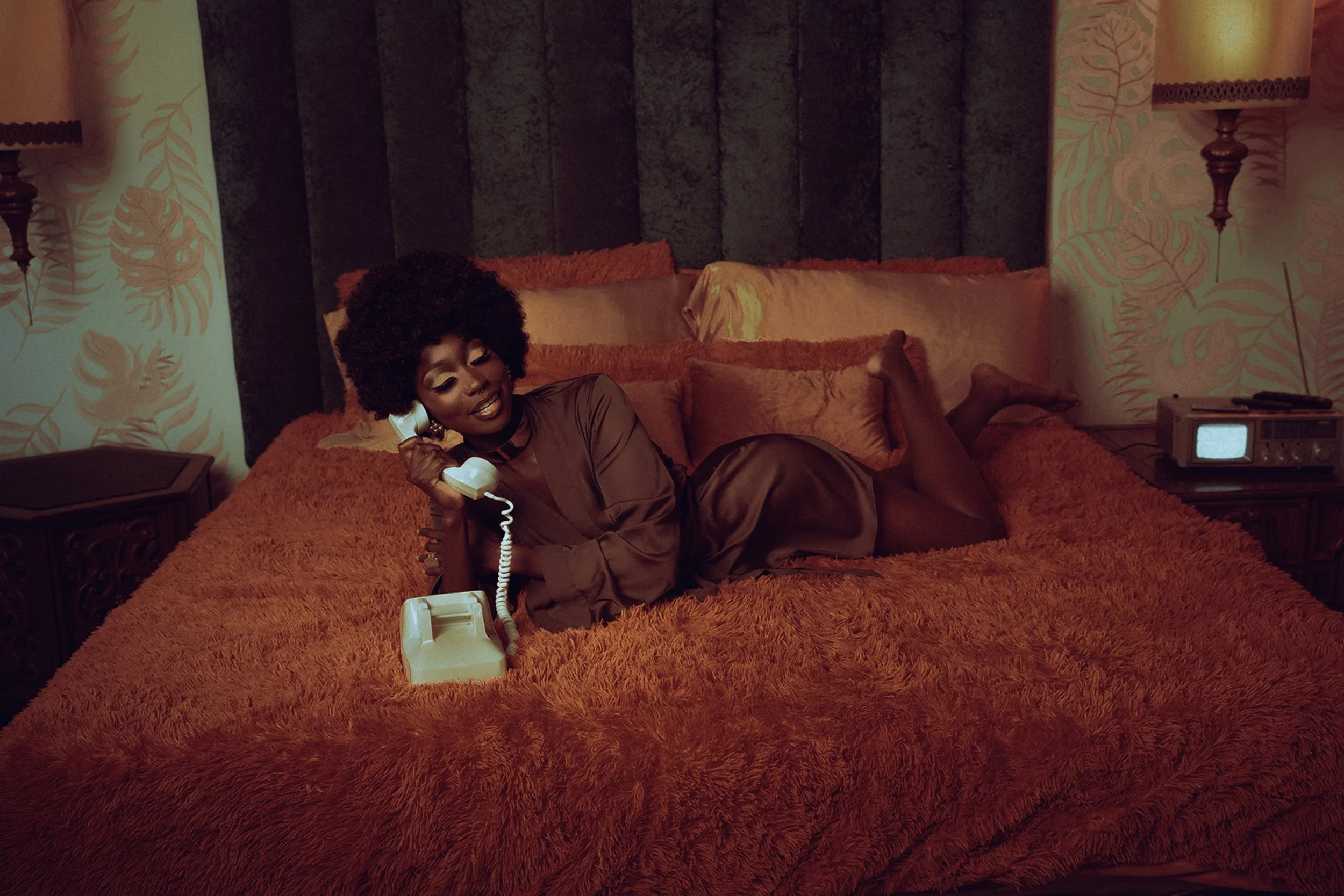 A woman with curly hair lying on a bed with orange bedding, talking on a vintage white rotary phone, in a retro-style bedroom with patterned wallpaper and lamps.