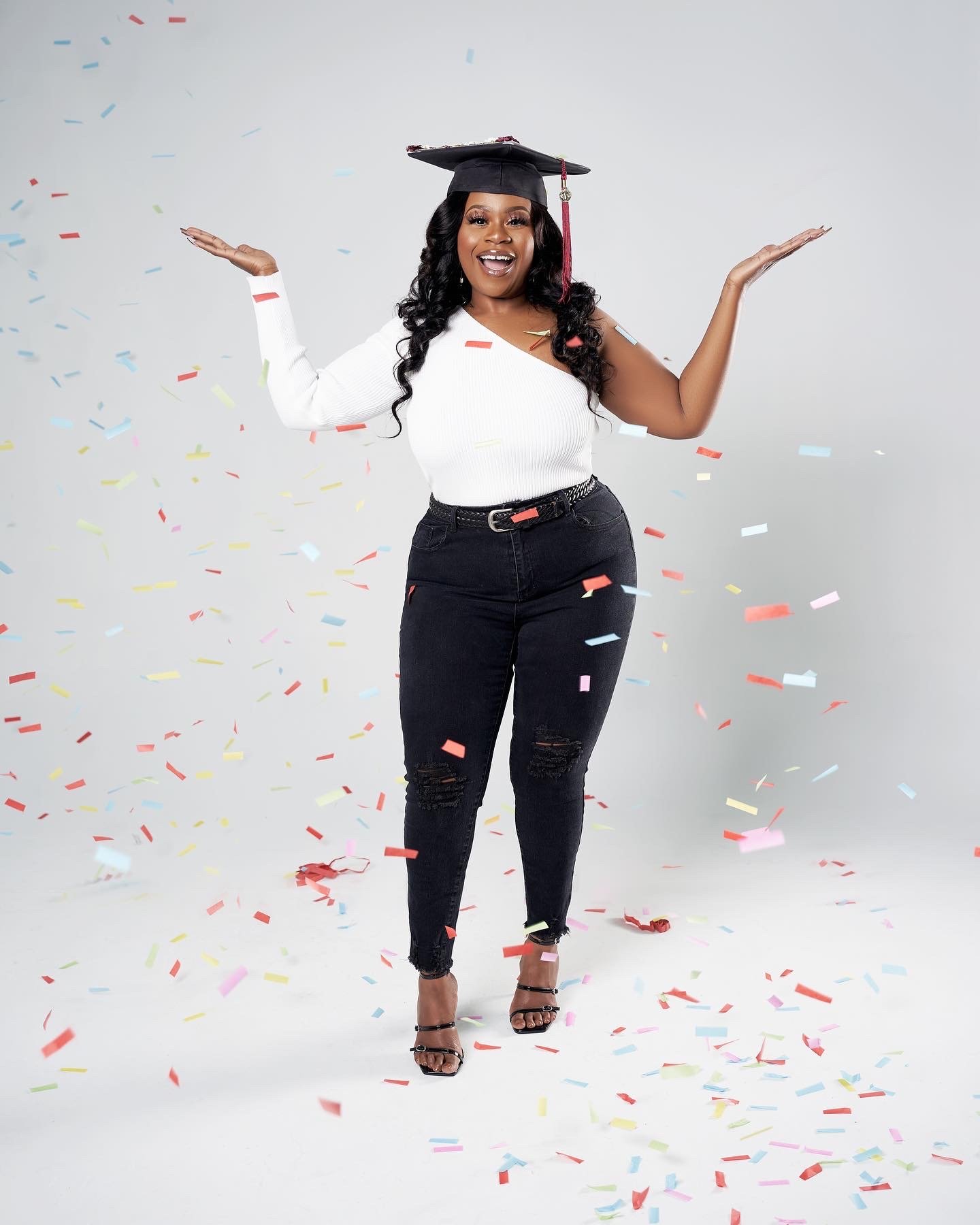 A woman in a white top and black ripped jeans celebrates with confetti falling around her, wearing a graduation cap.