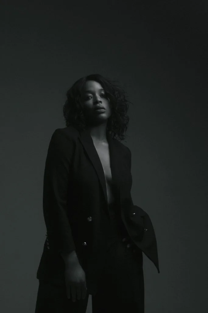 A woman with curly hair wearing a black blazer and black pants stands against a plain dark background.