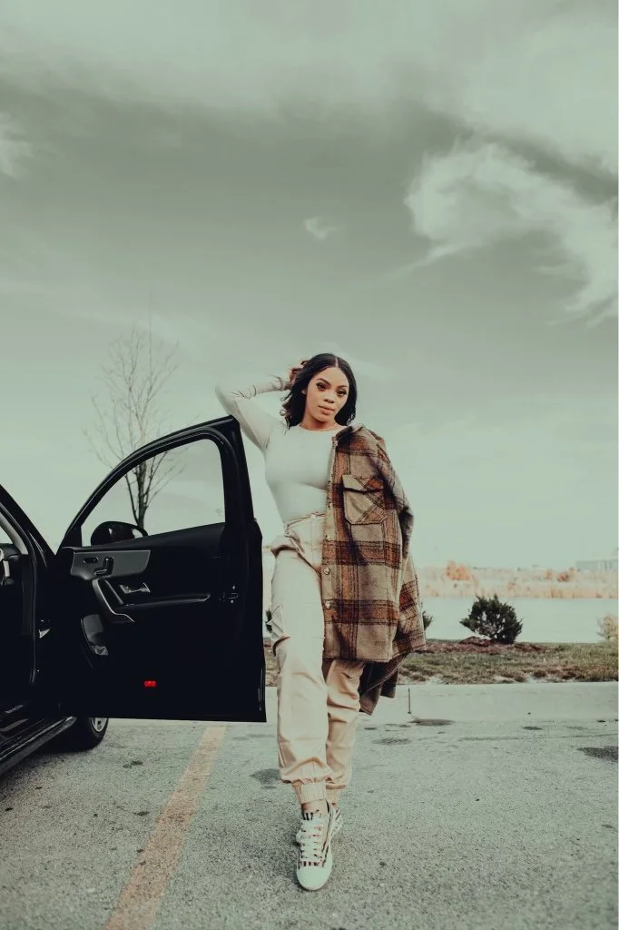 Young woman standing next to a black car with the driver's door open, near a body of water with cloudy sky in the background.