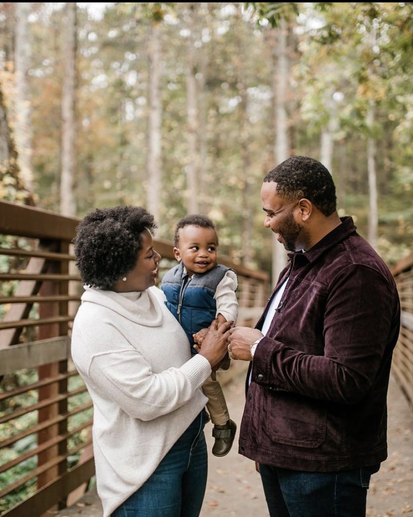A family of three — a woman, a man, and a young boy — standing on a wooden bridge in a forest, smiling and enjoying a moment together.