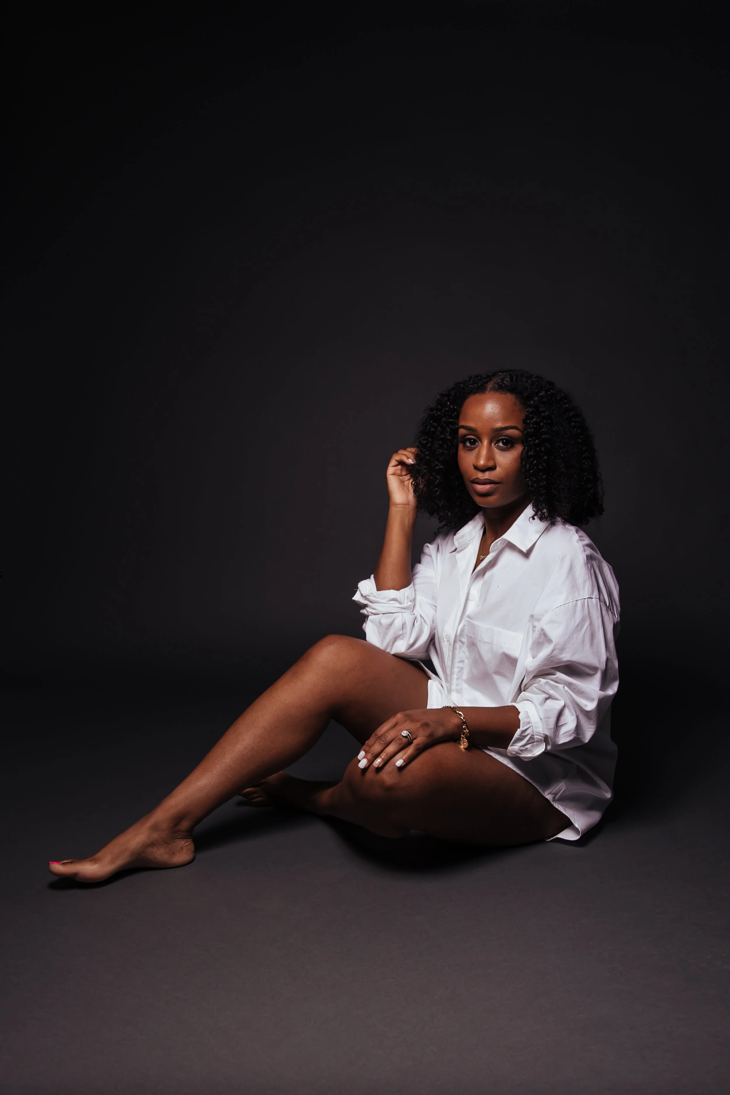 A woman with curly hair, wearing a white shirt, sitting on the floor against a dark background.