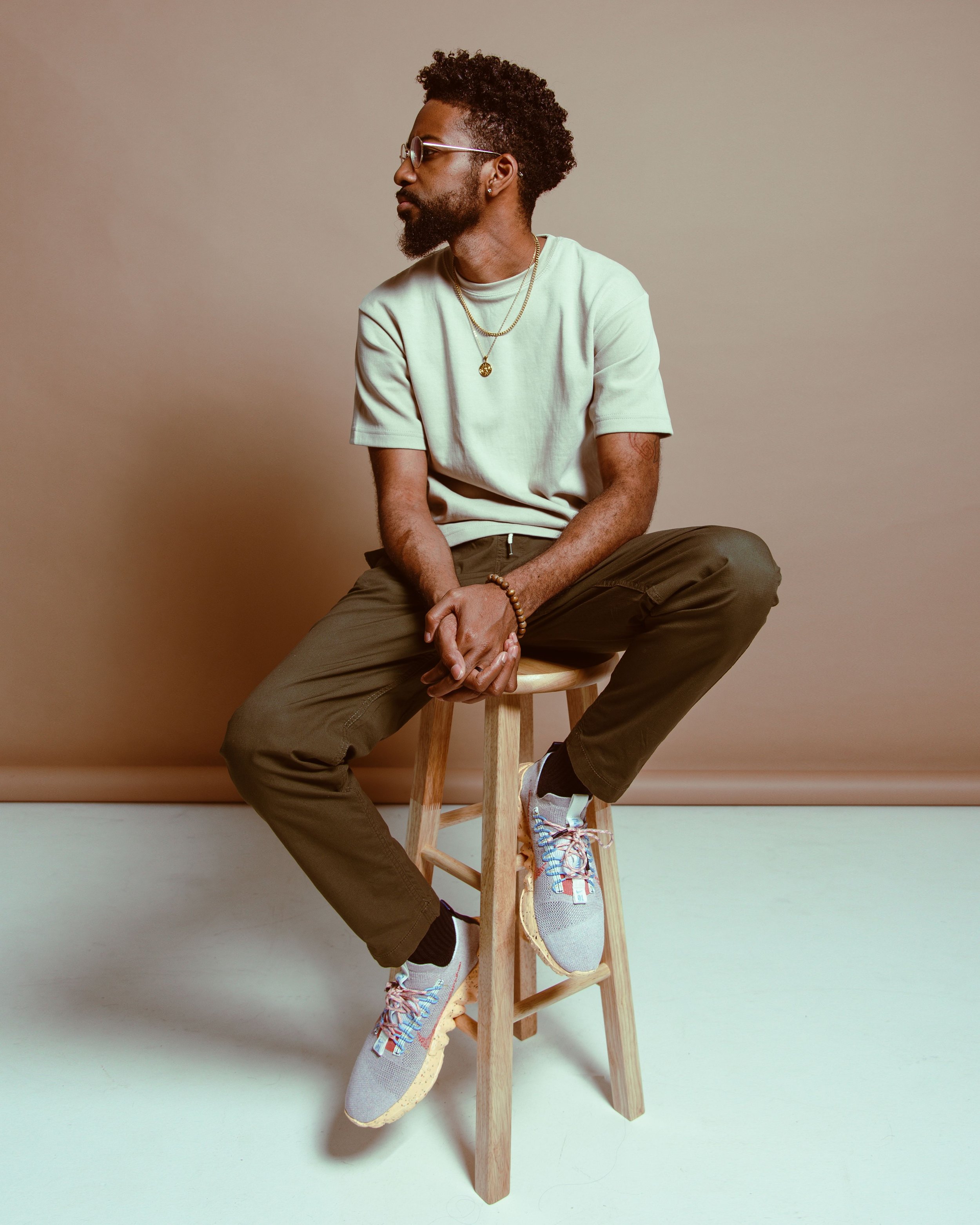 A man with curly hair, beard, sunglasses, gold chains, and a bracelet, wearing a light-colored t-shirt, brown pants, and sneakers, sitting on a wooden stool against a beige backdrop.