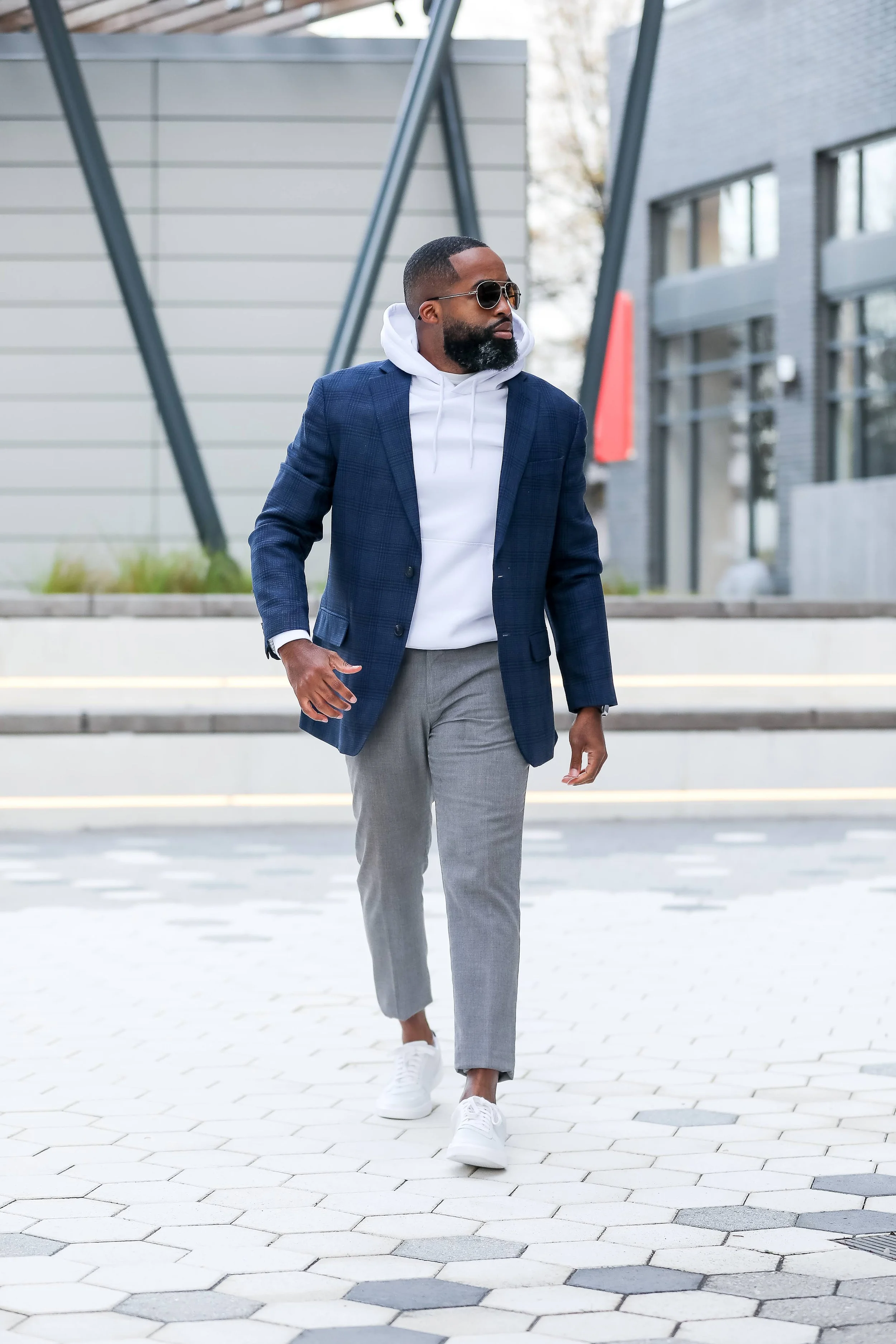 A man walking outside wearing sunglasses, a white hoodie, a blue blazer, gray pants, and white sneakers.