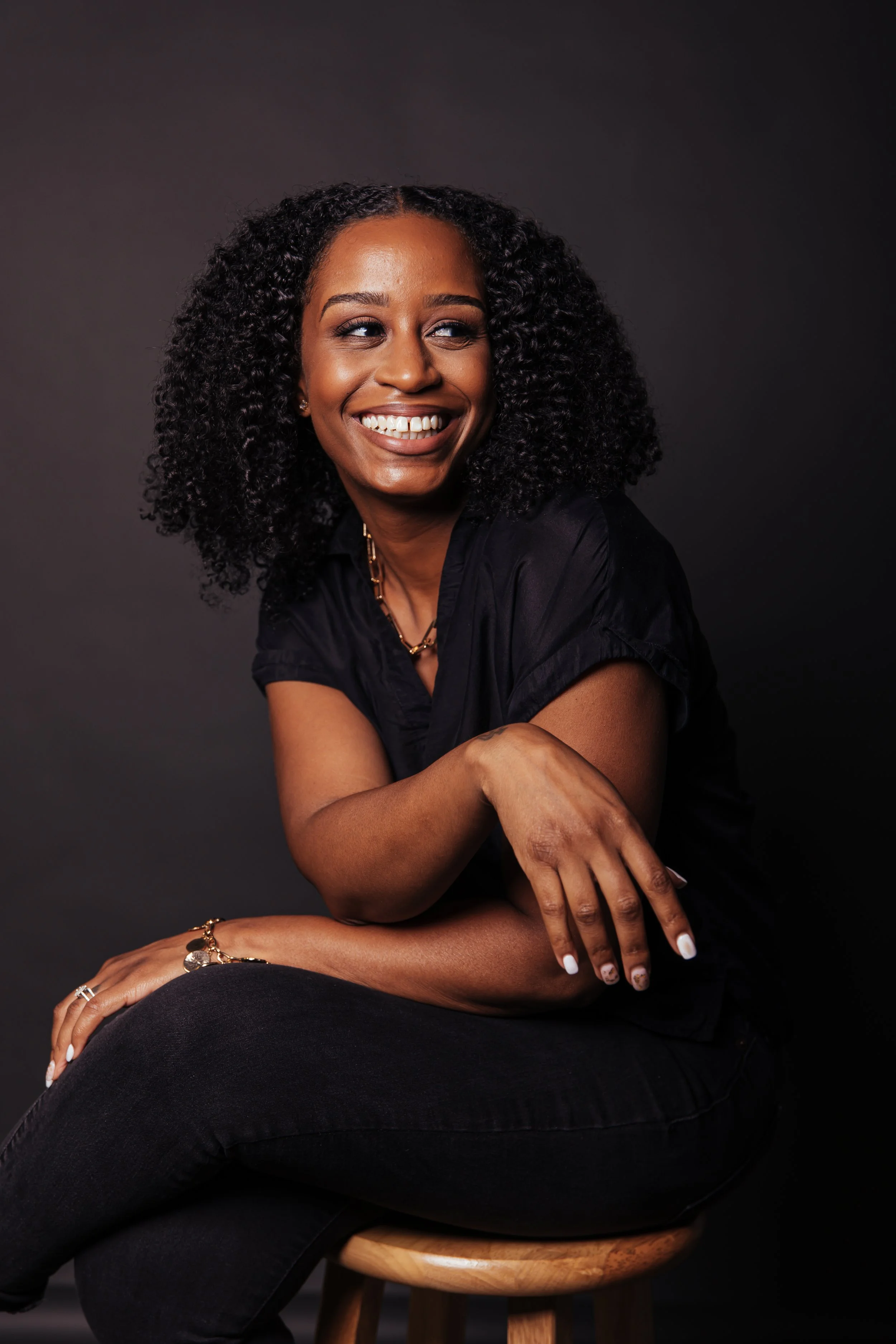 A woman with curly black hair smiling and looking to her right, wearing a black shirt and black jeans, sitting on a wooden stool against a dark background.