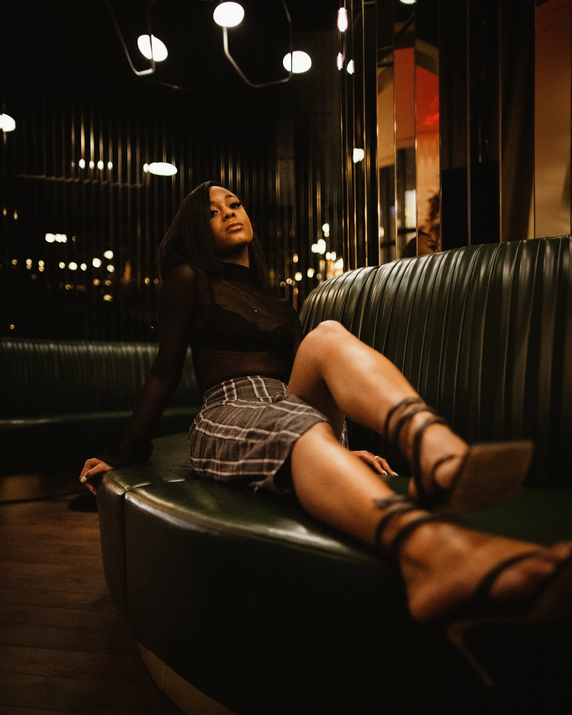 A woman sitting on a green leather bench in a dimly lit setting with hanging lights overhead, wearing a black sheer top, plaid skirt, and high-heeled shoes