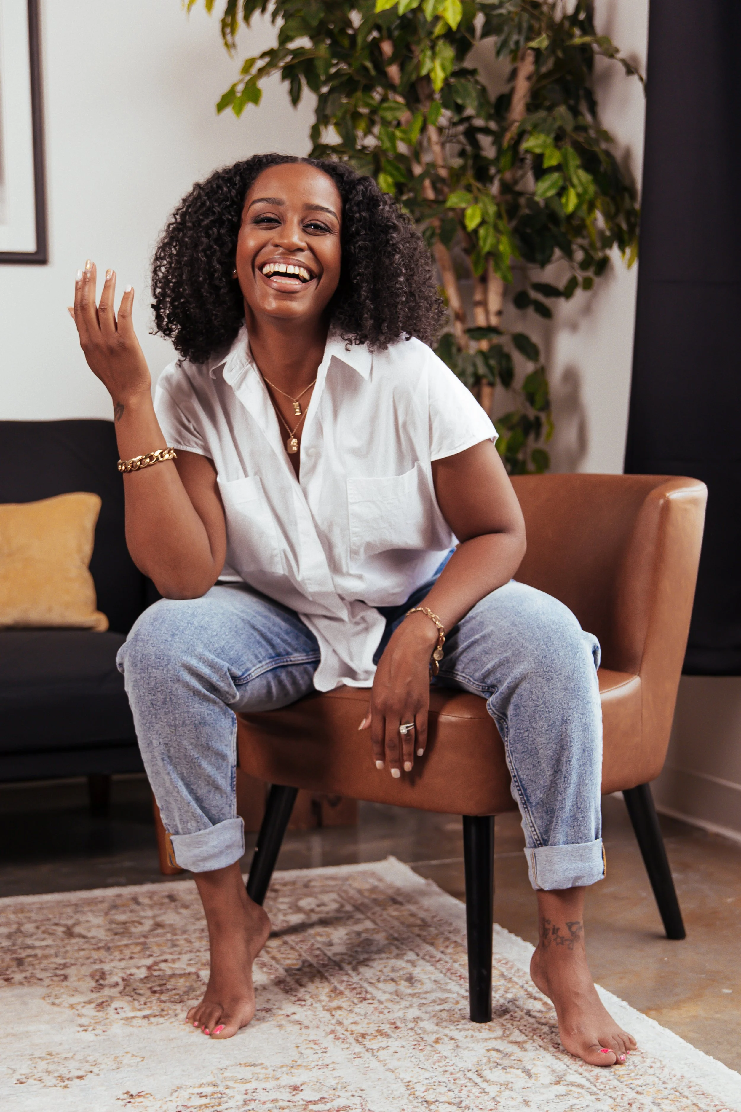 A smiling woman with curly hair, wearing a white shirt and jeans, sitting barefoot on a tan chair in a cozy room with a black sofa, yellow pillow, large green plant, and a patterned rug.