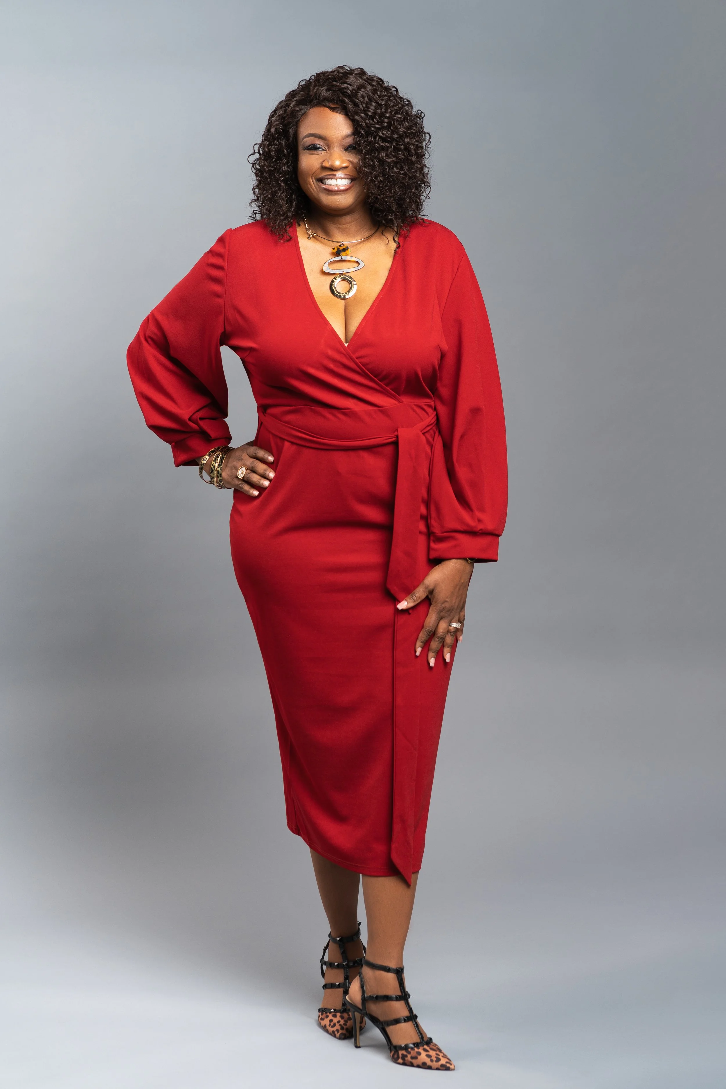 A woman in a red dress stands smiling against a gray background, wearing leopard print heels and jewelry.