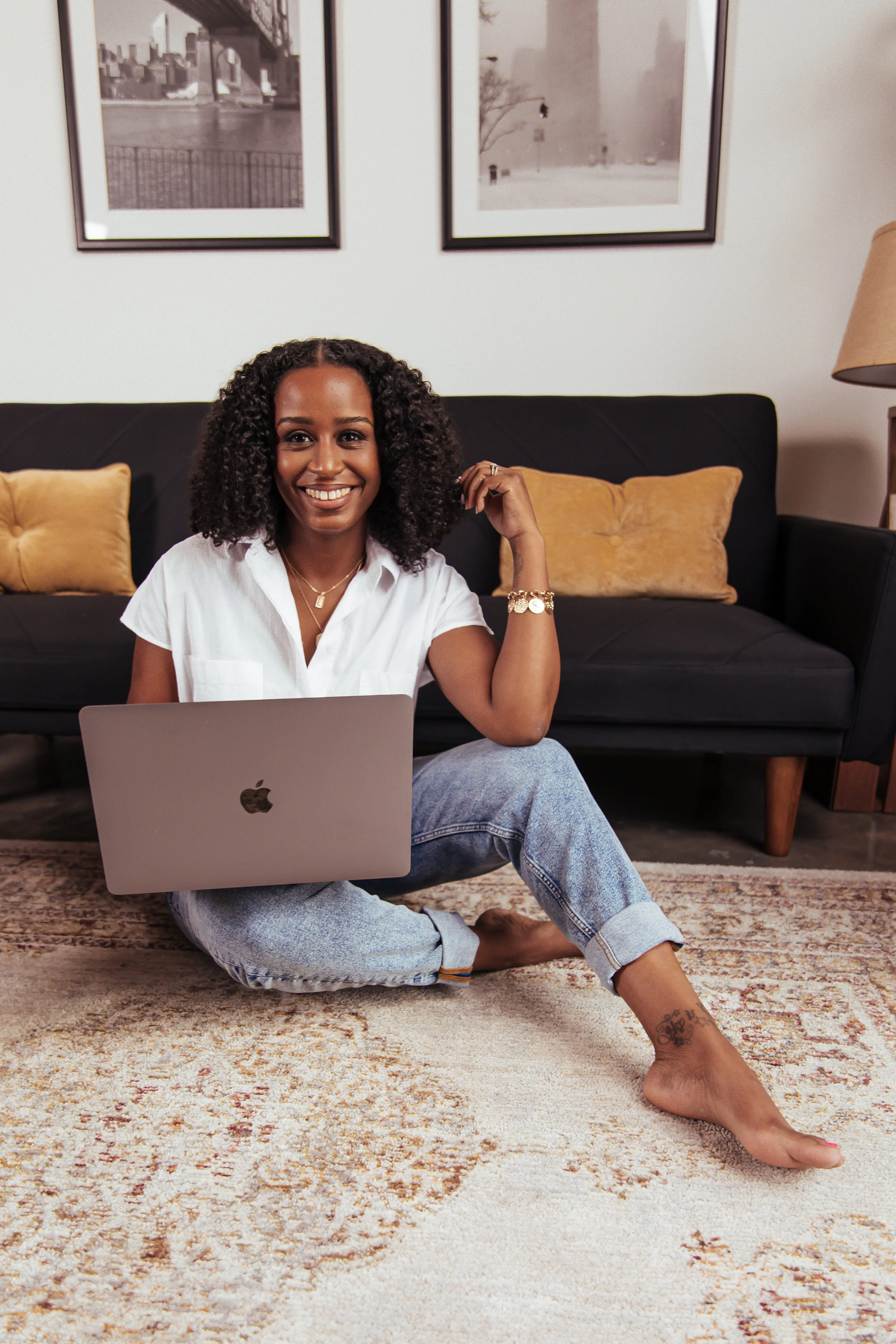 A woman sitting on a rug in a living room with a laptop, smiling at the camera, with a black sofa and framed pictures in the background.