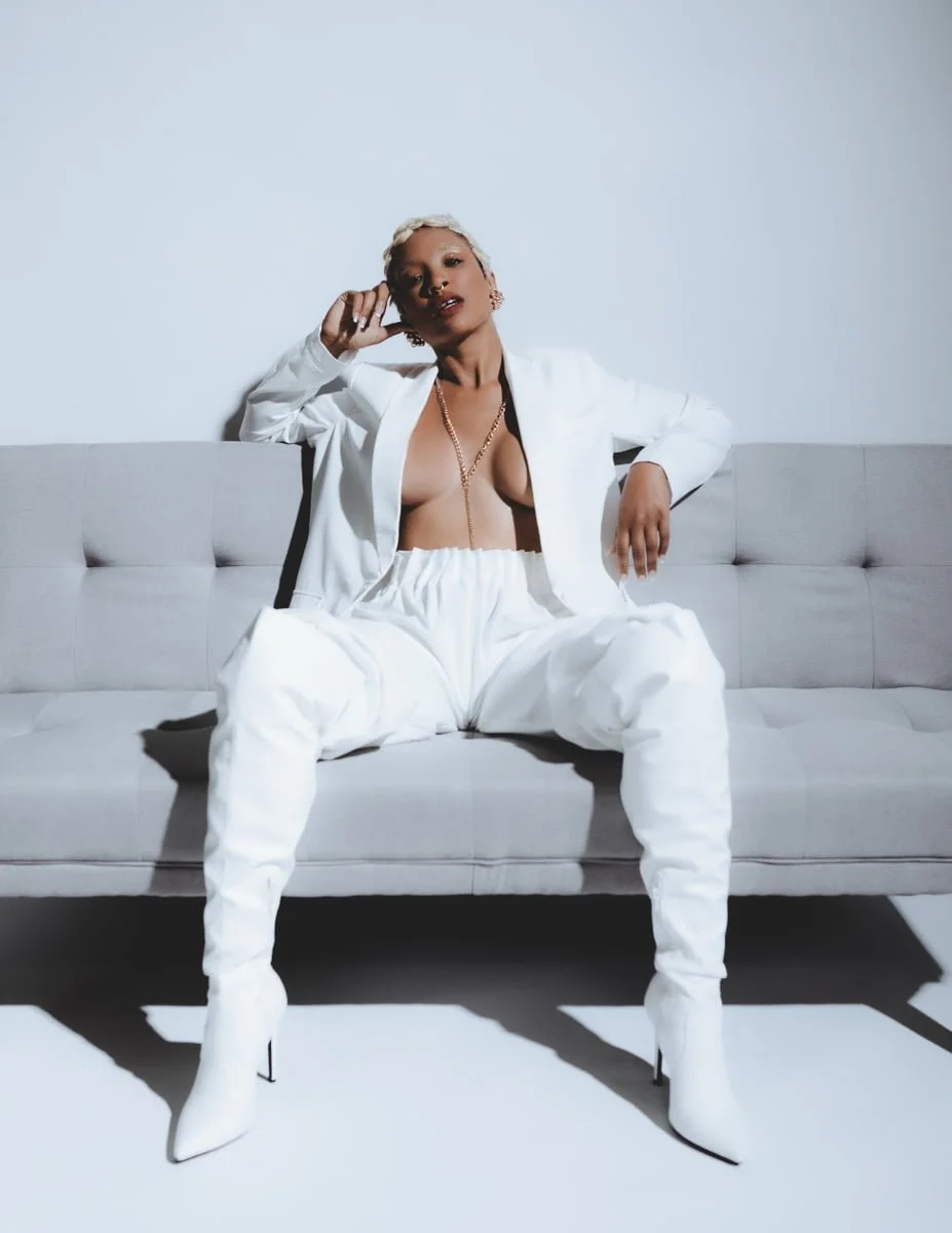 A woman with blonde hair sitting on a gray sofa, wearing a white blazer, white baggy pants, white high-heeled boots, and jewelry, posing with a confident expression in a minimalist studio setting.