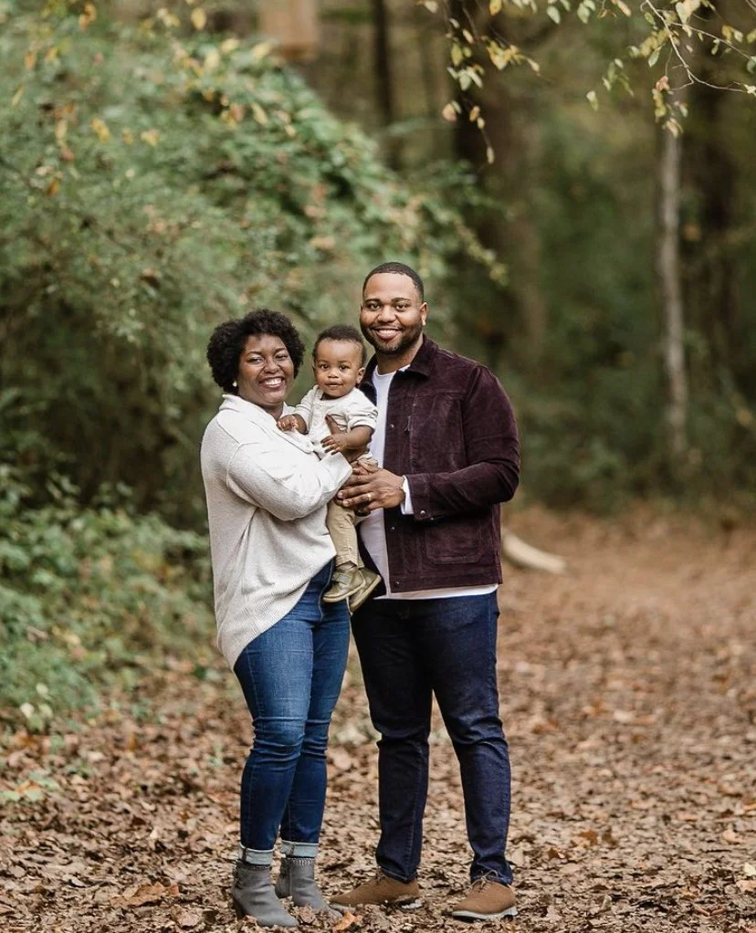 Happy family of three standing on a wooded path with autumn leaves, smiling at the camera.