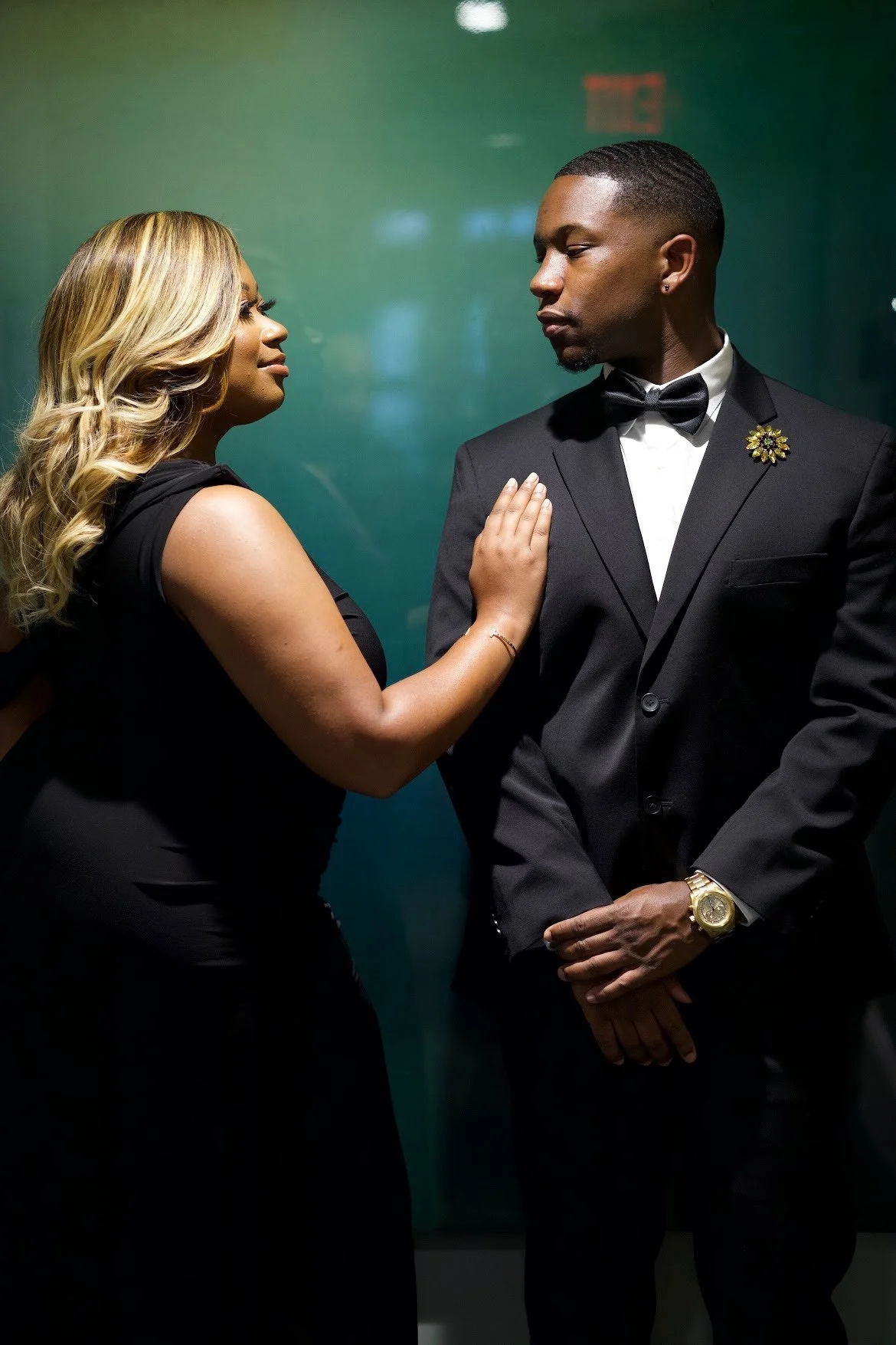 A woman in a black dress touching the shoulder of a man in a tuxedo, both looking at each other, in front of a dark green background.