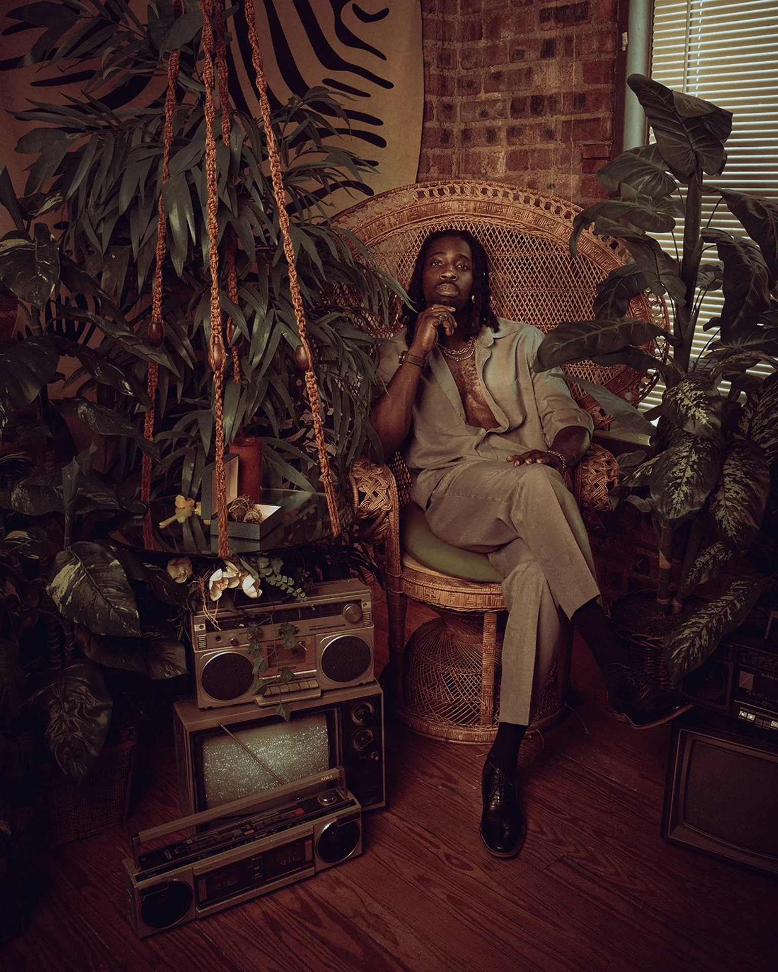 A man sitting on a wicker peacock chair surrounded by large indoor plants, with a retro boombox on the floor nearby, in a room with exposed brick wall and window blinds.