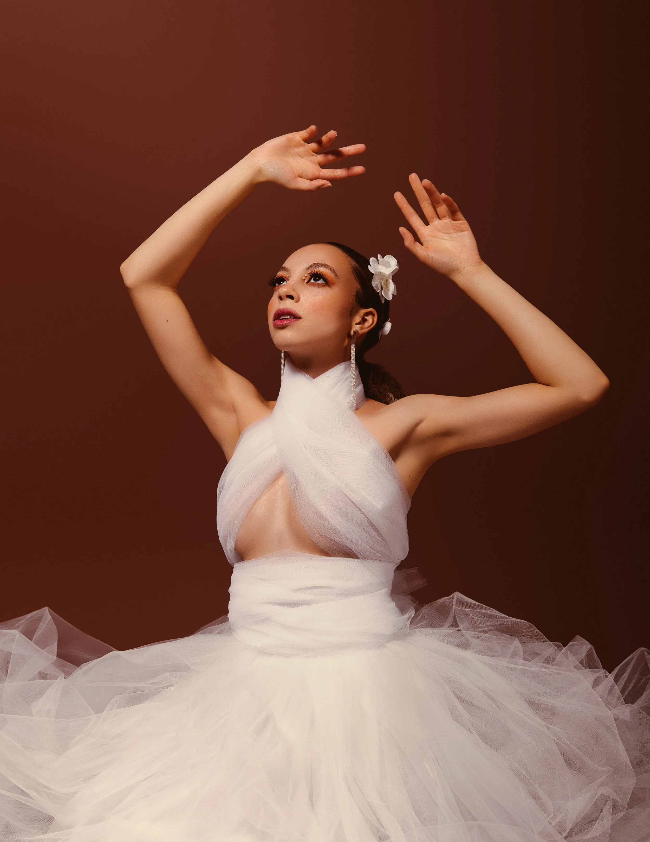 A woman in a white ballet costume with tulle fabric, floral hair accessory, and makeup, posed with arms raised against a plain background.