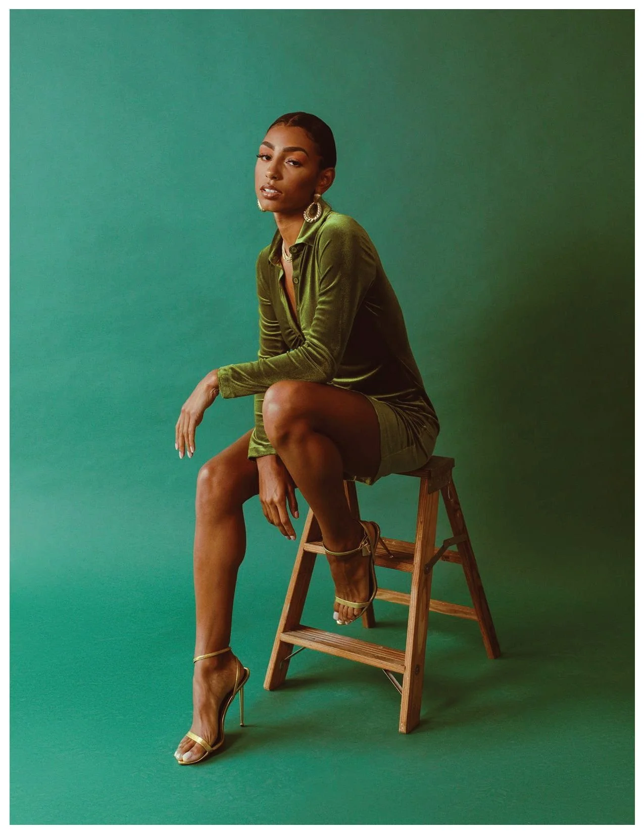 A woman with dark hair in a sleek style, wearing an olive-green velvet shirt and matching shorts, gold heels, and large earrings, seated on a wooden step ladder against a green background.
