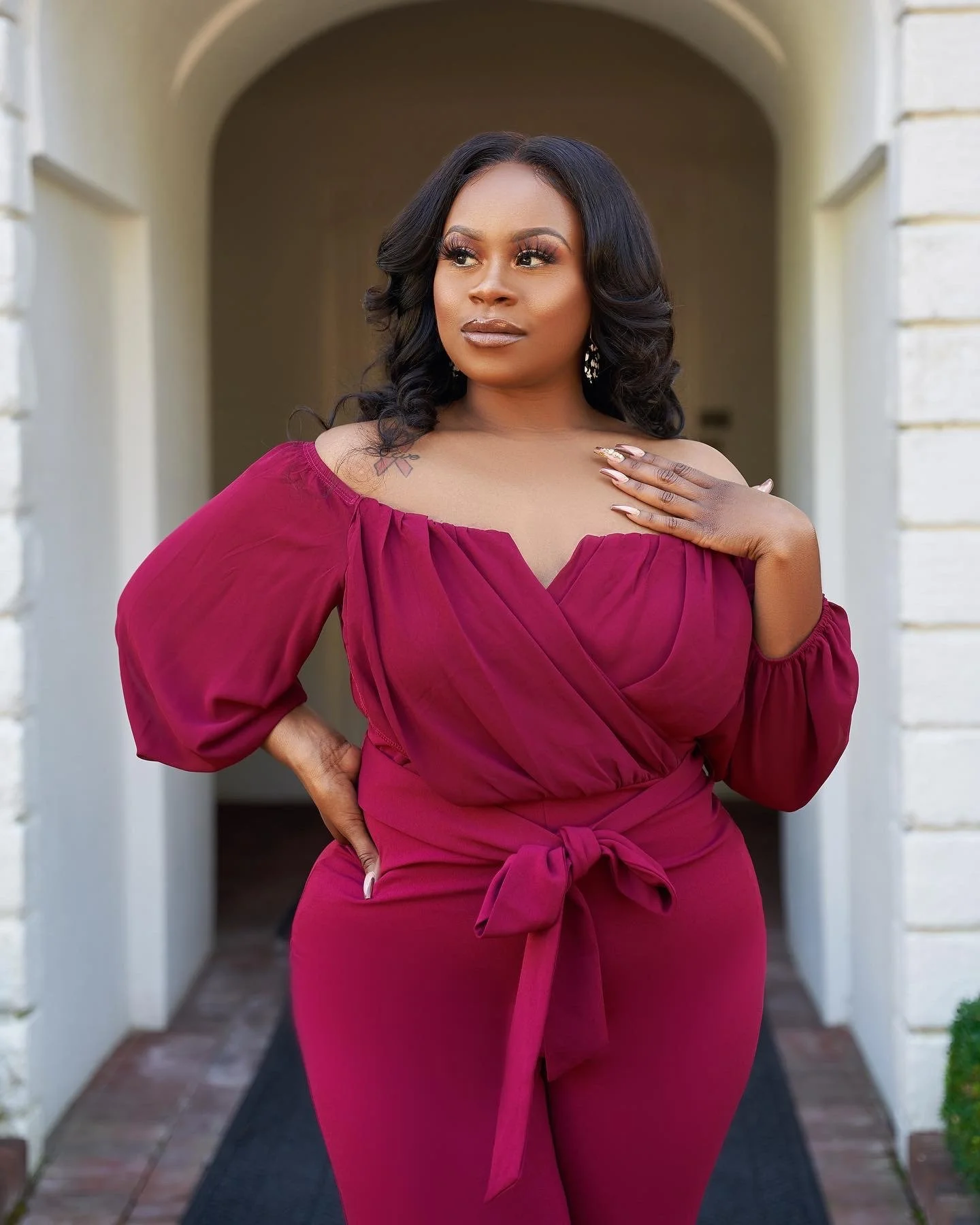 A woman in a stylish, off-shoulder maroon dress with puffed sleeves, posing confidently in front of a white arched entrance.