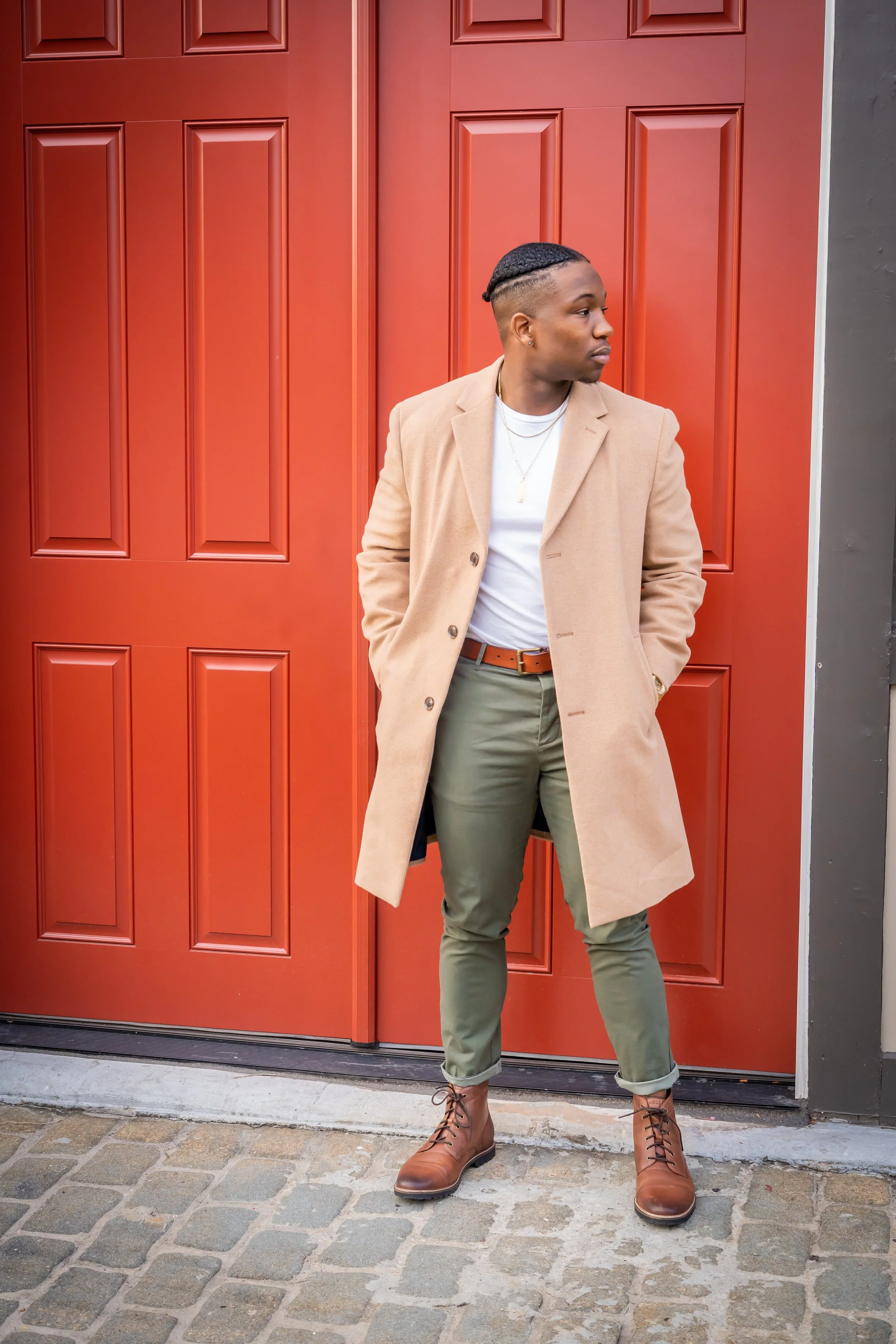 A young man standing in front of a red door, wearing a beige coat, white t-shirt, olive green pants, and brown boots, looking to his right with his hands in his pockets.