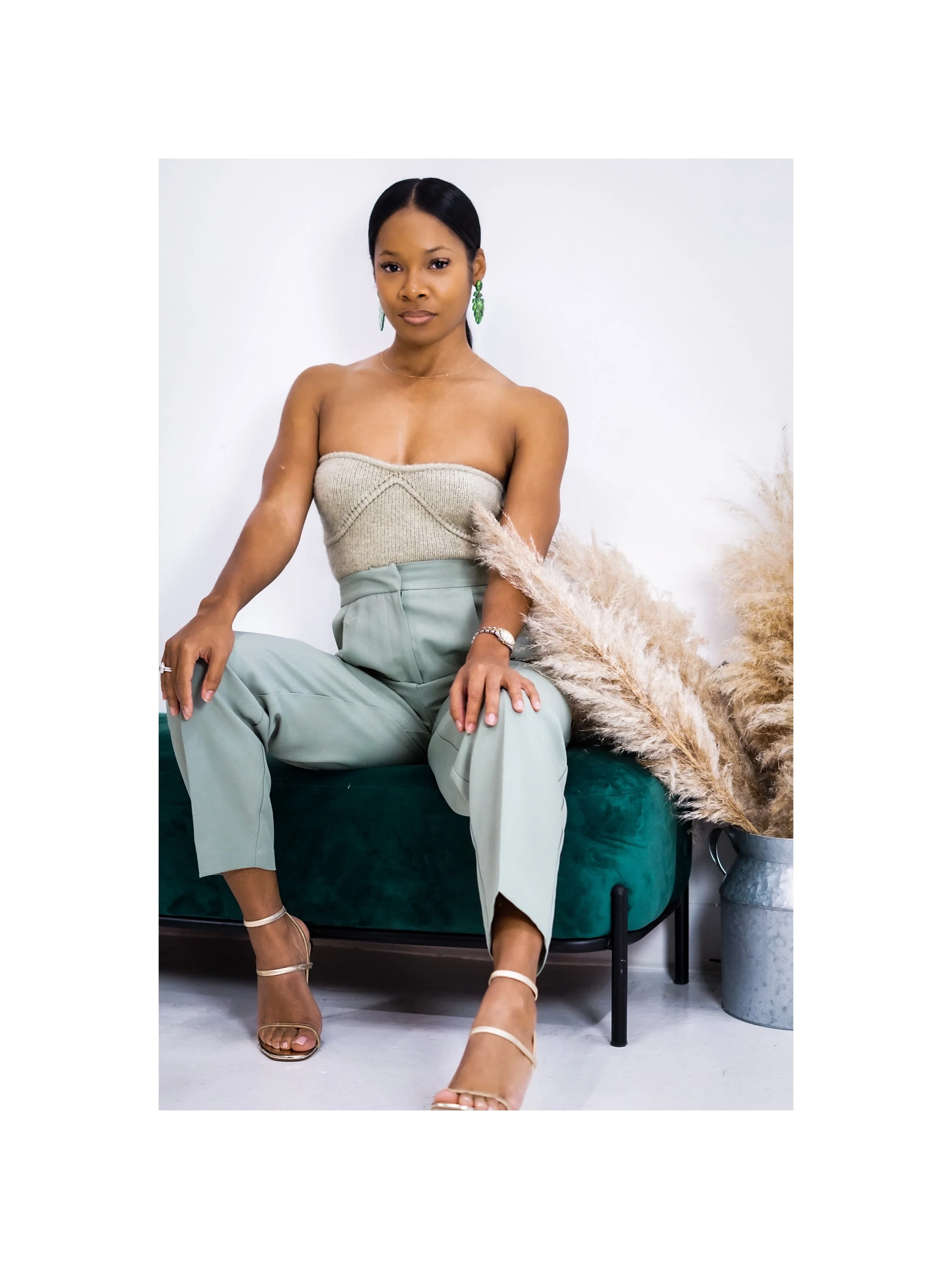 A woman sitting on a green velvet bench, wearing a beige strapless top, high-waisted light green pants, and nude high heels, with pampas grass in a pot beside her.