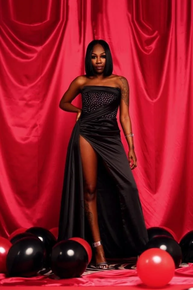 A woman standing in front of red curtains, wearing a black strapless dress with a high slit, revealing her leg and wearing high heels, surrounded by black and red balloons.