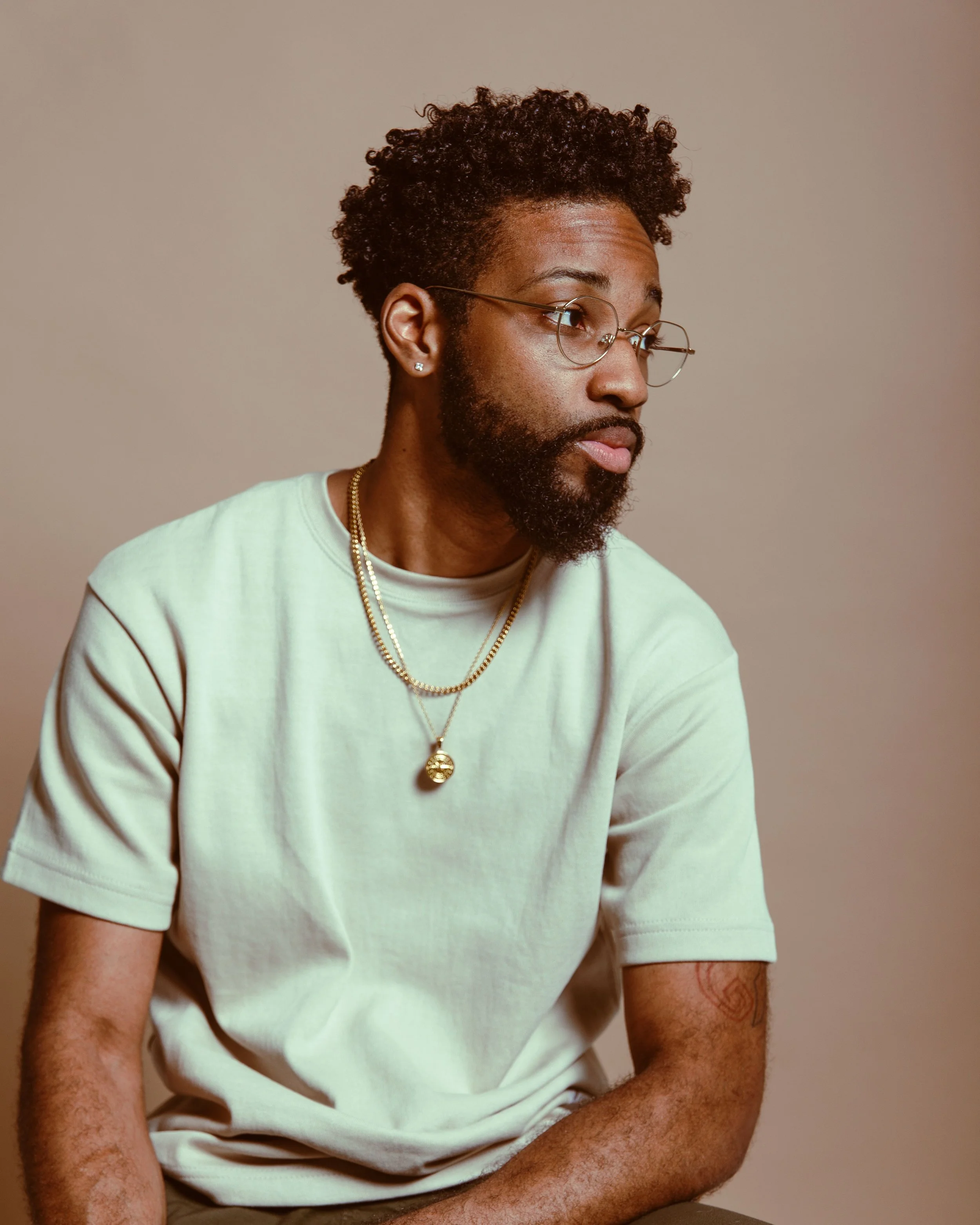 A young man with a beard and glasses, wearing a beige t-shirt and gold jewelry, looking to the right against a plain background.