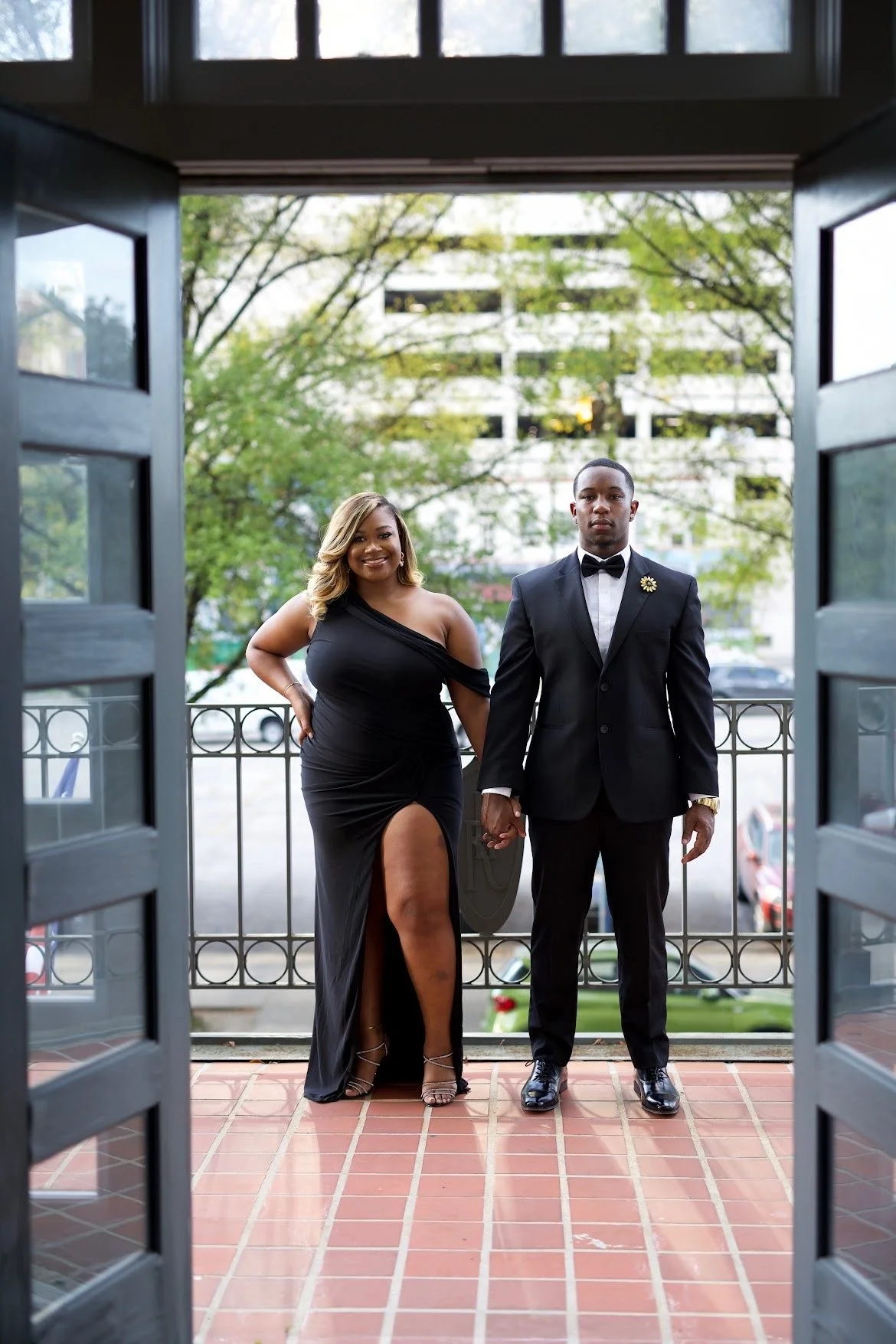 A couple holding hands on a balcony, dressed formally for a special occasion, with a cityscape and trees in the background.