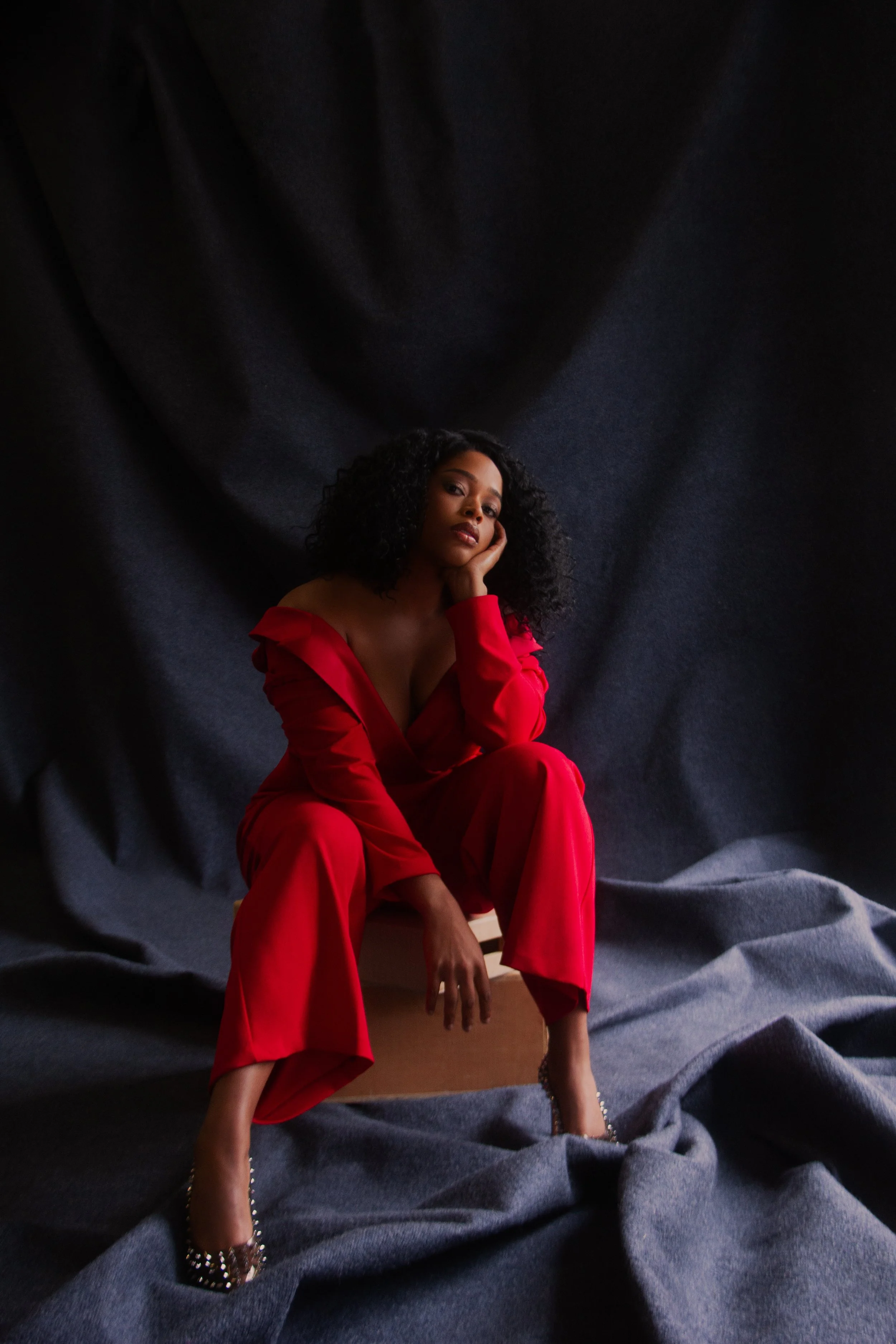 A woman with curly black hair, wearing a red off-shoulder outfit and patterned high heels, sitting on a wooden stool against a dark fabric backdrop, with her hand resting on her face.