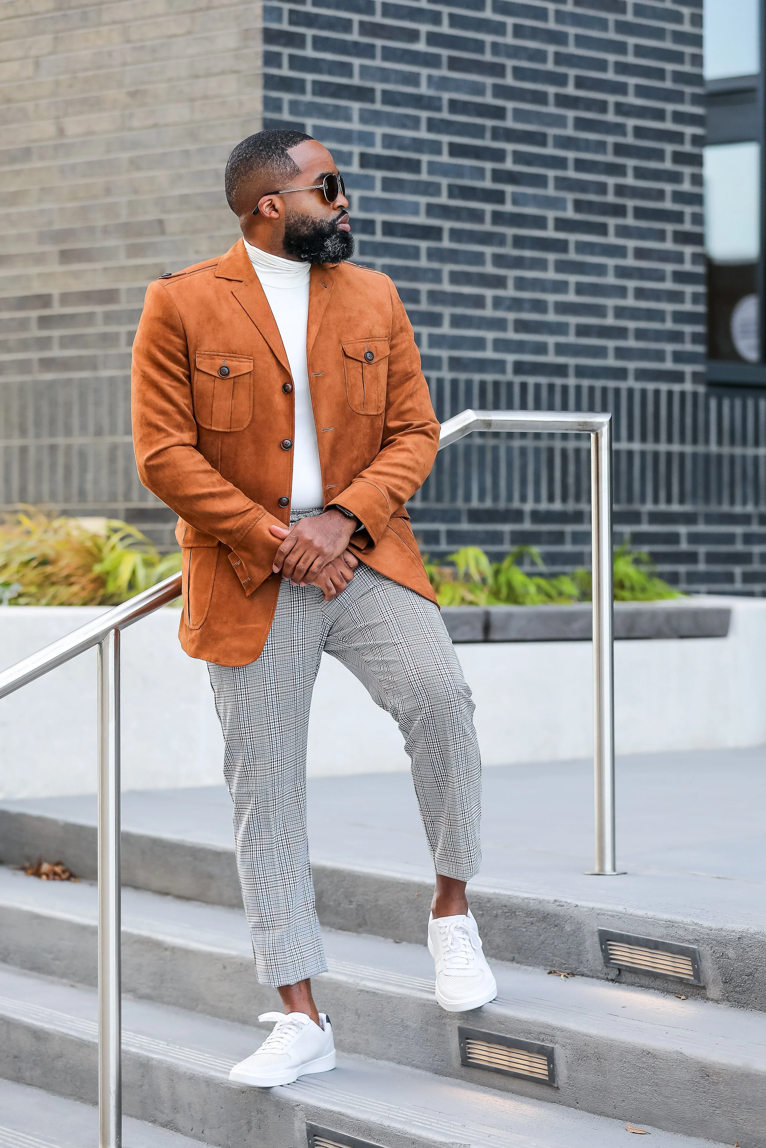 A man with a beard, wearing sunglasses, a white turtleneck, a brown suede blazer, checkered pants, and white sneakers, standing on outdoor concrete stairs near a black brick building.