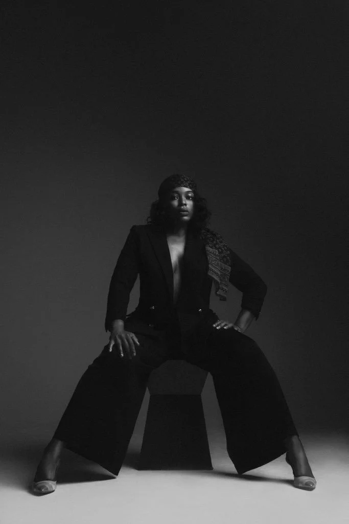 Black and white portrait of a woman sitting on a stool, posed with legs wide apart, in a studio setting with a plain dark background.