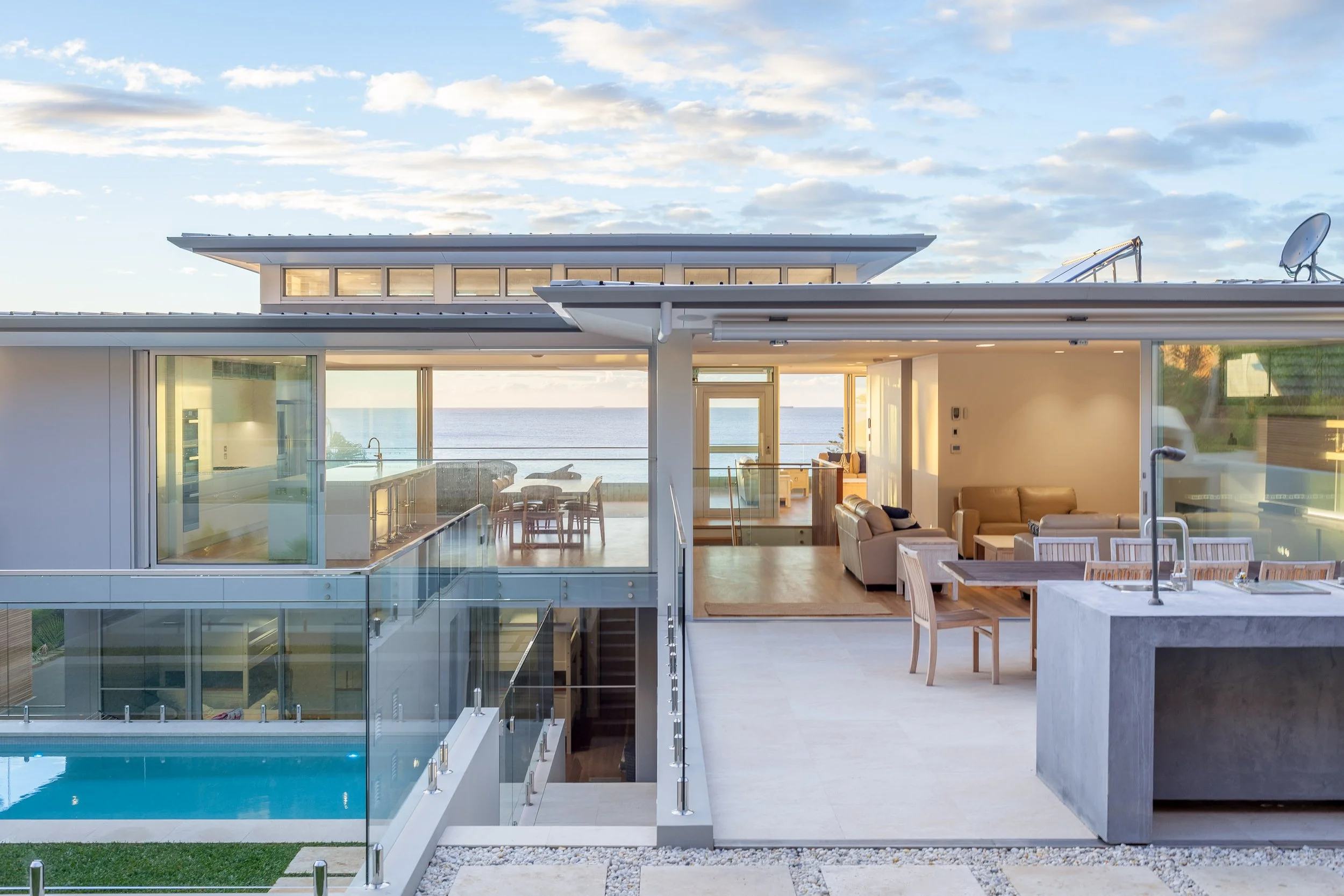 Modern house with large glass windows and doors, balcony with outdoor furniture, ocean view in the background, interior with living area and kitchen, outdoor pool area with stairs.