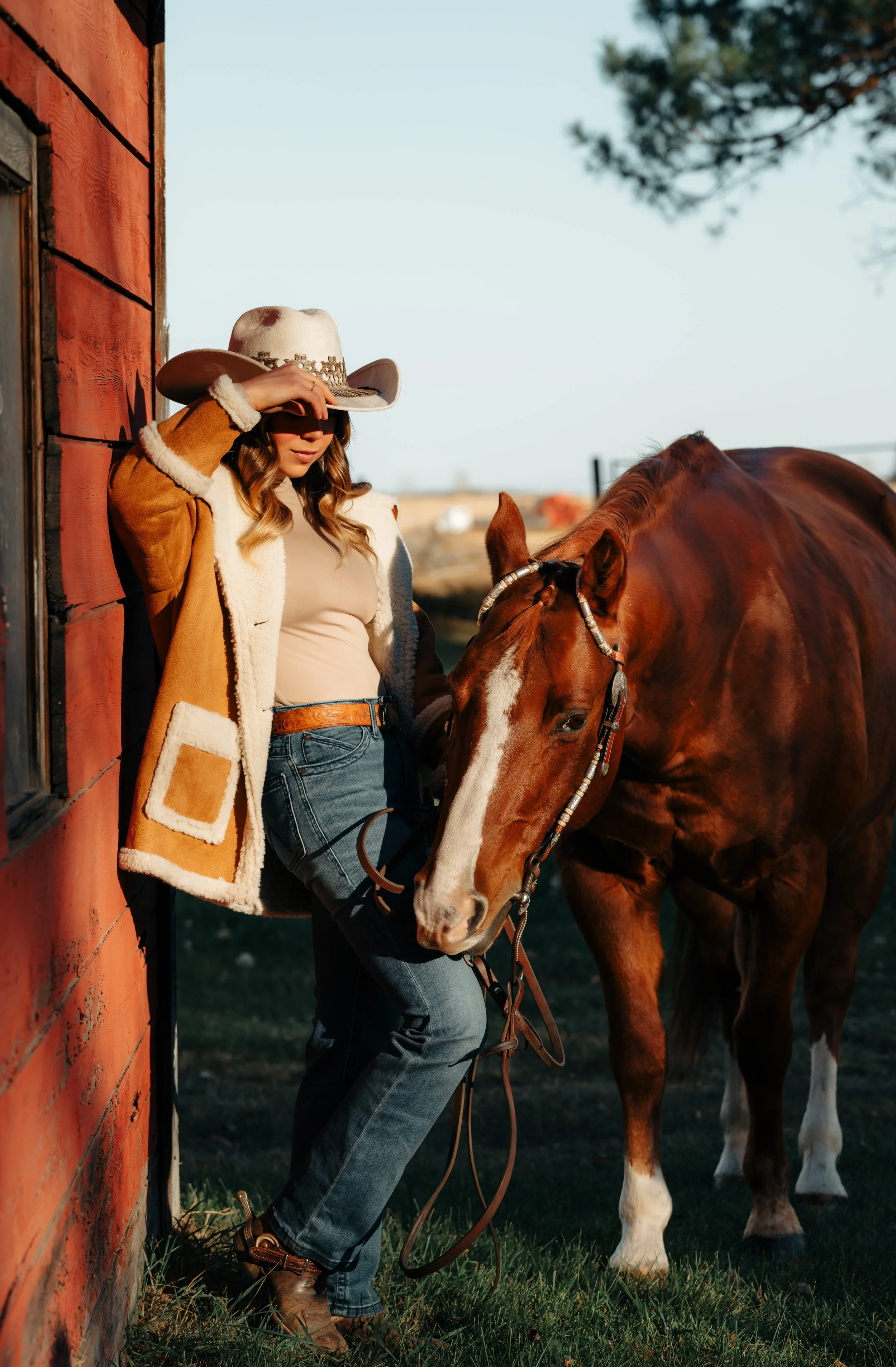 Western woman in shearling coat and cowboy hat standing beside her sorrel horse against a red barn in Alberta