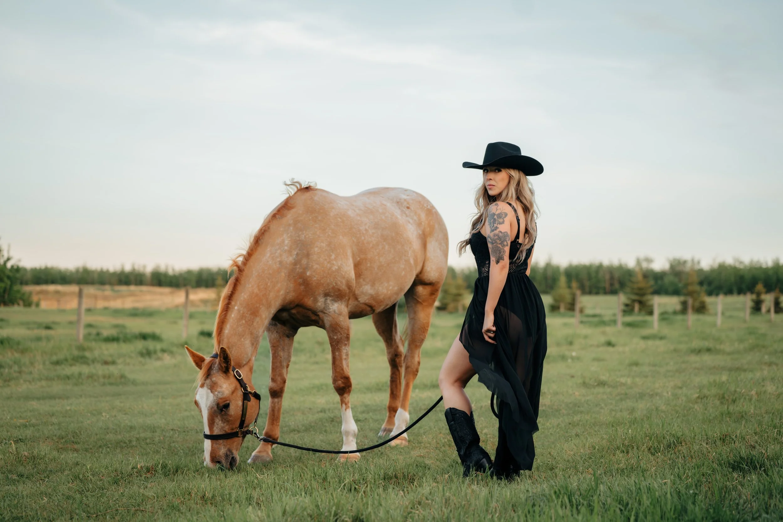 Tattooed cowgirl in black dress and wide-brimmed hat standing beside her grazing appy in Alberta field